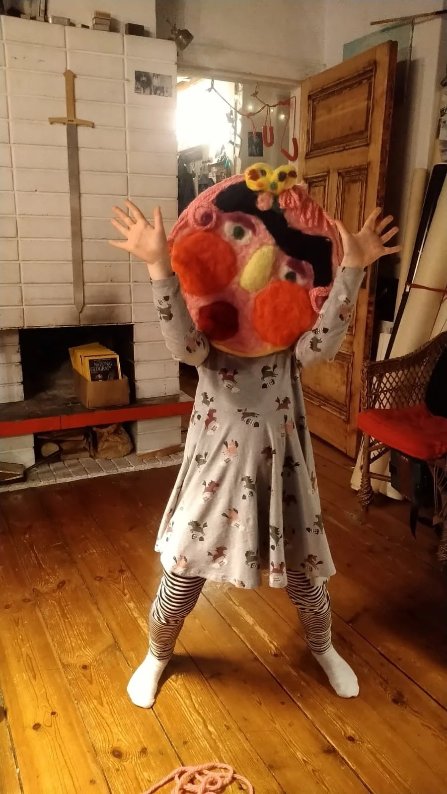 Child wearing a large, colorful, plush mask resembling a face with red cheeks, a yellow nose, black hair, and small yellow fluffy accessories on top, dressed in a gray dress with animal patterns and striped leggings, standing in a cozy living room wi