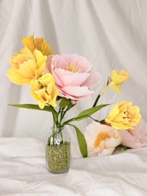 A glass vase holding a bouquet of large pink and yellow artificial flowers with green leaves, placed on a white fabric surface with a white fabric backdrop.