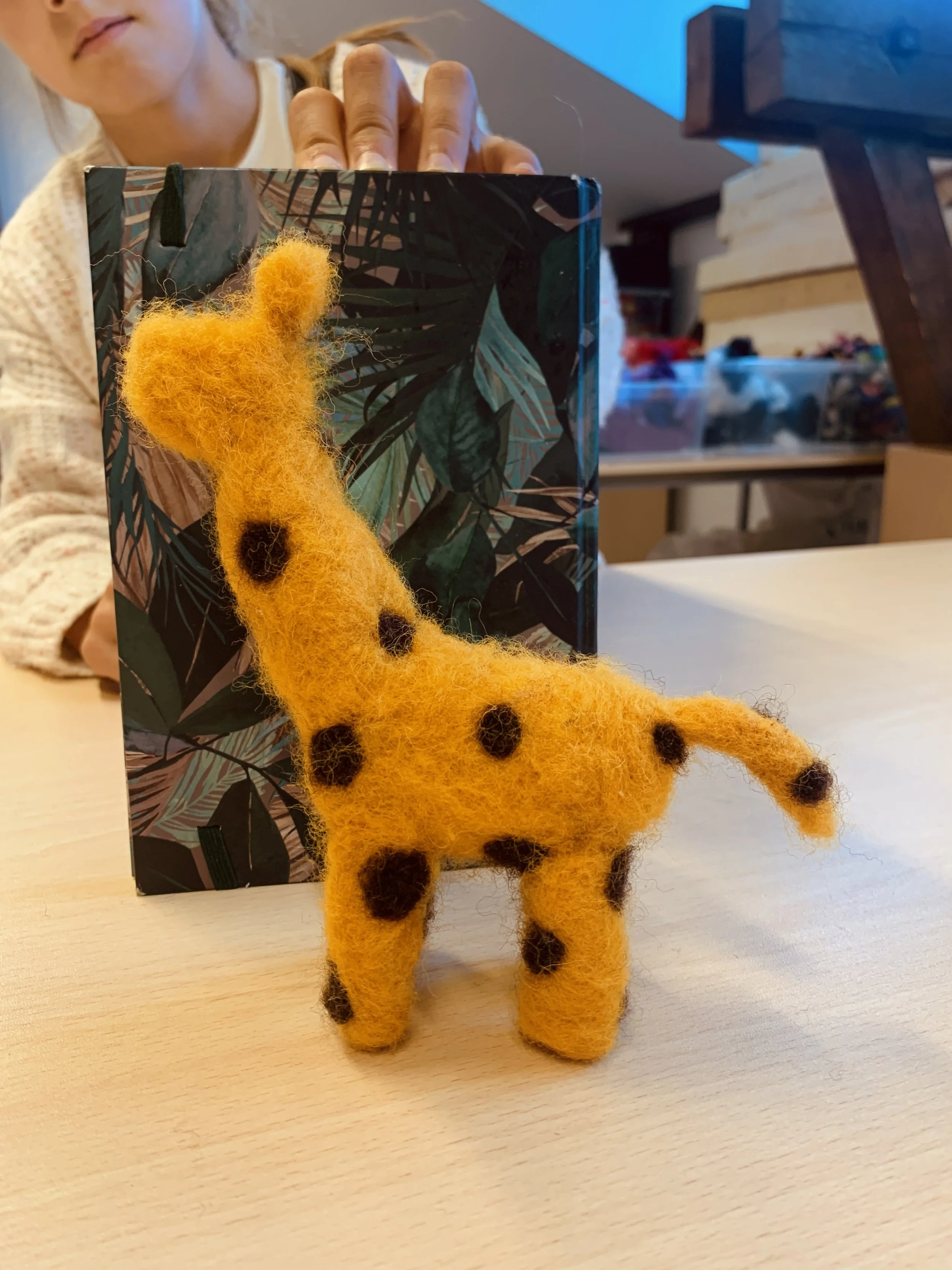A yellow felt giraffe with brown spots sitting on a light-colored wooden surface, with a partly blurred person in the background holding a decorative object with a tropical leaf pattern.