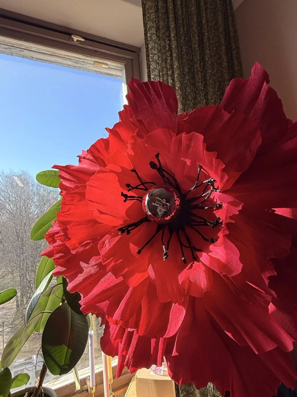 Close-up of a large red paper flower decoration with black wire stamens, placed near a window with a view of trees and a blue sky outside.