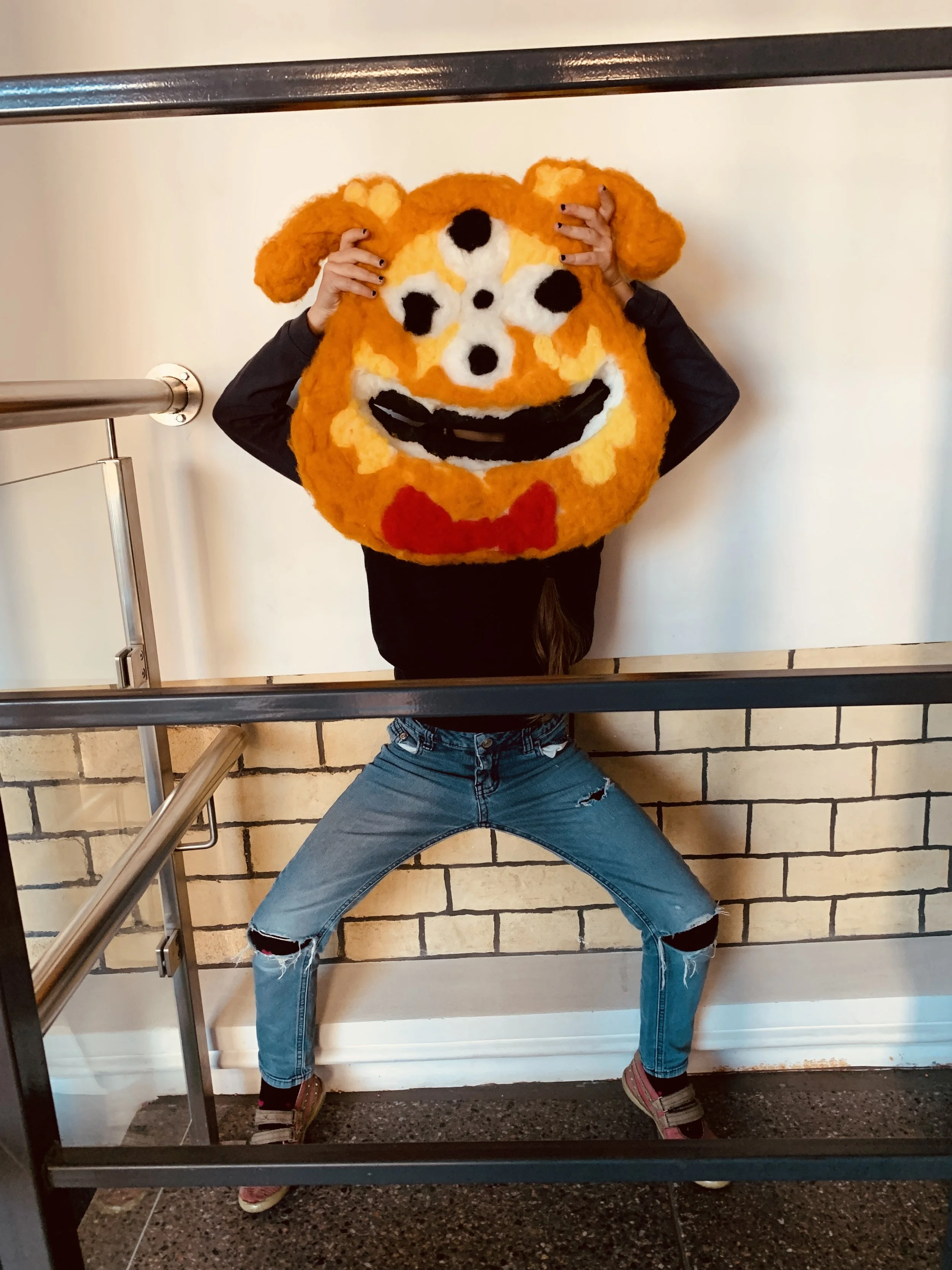 Person holding a large plush emoji mask with a smiling face, dog ears, and a red bowtie, standing behind a black metal railing against a brick wall background.