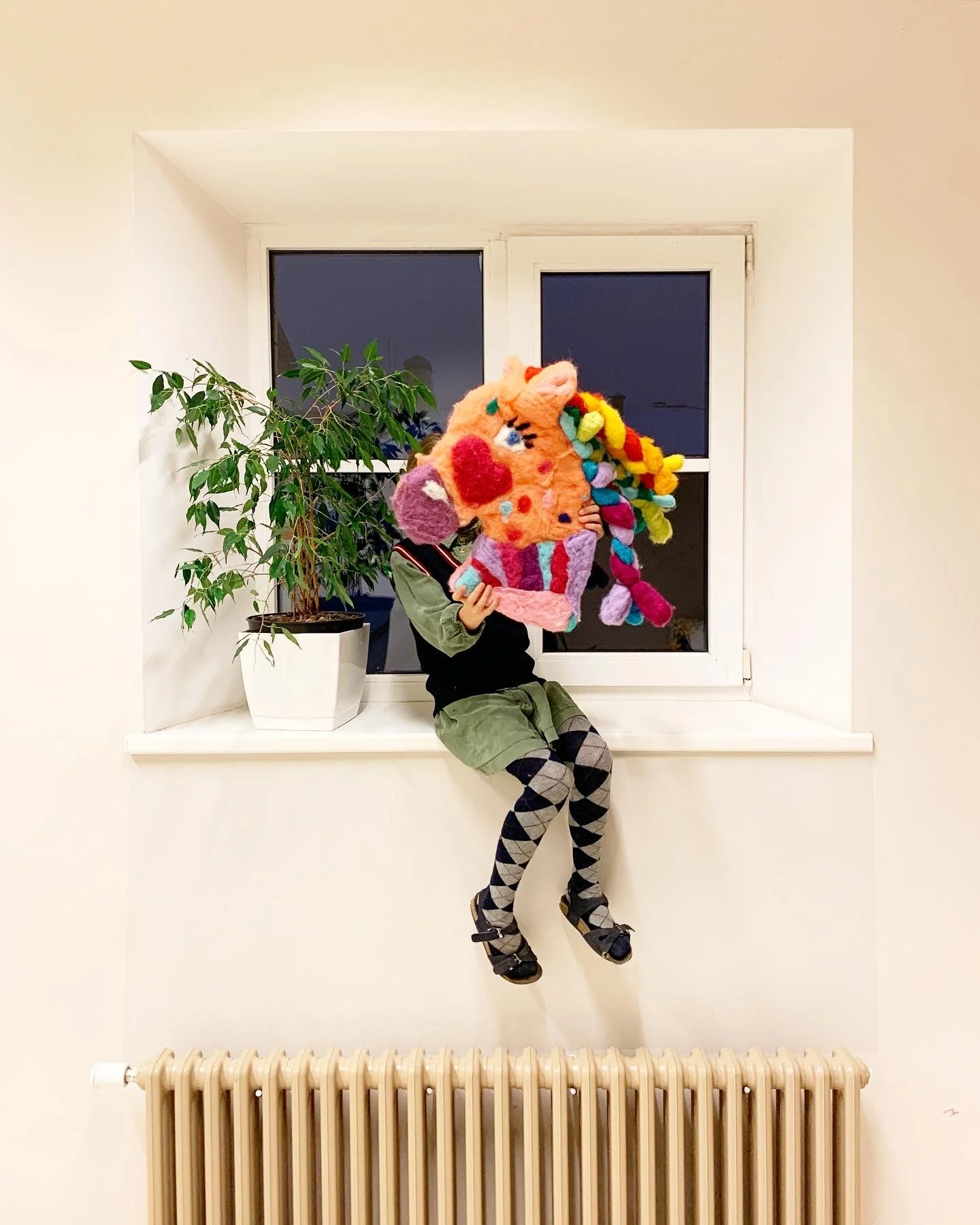 A person sitting on a windowsill holding a large colorful plush toy in front of their face, with a potted plant on the window ledge and a dark window behind.