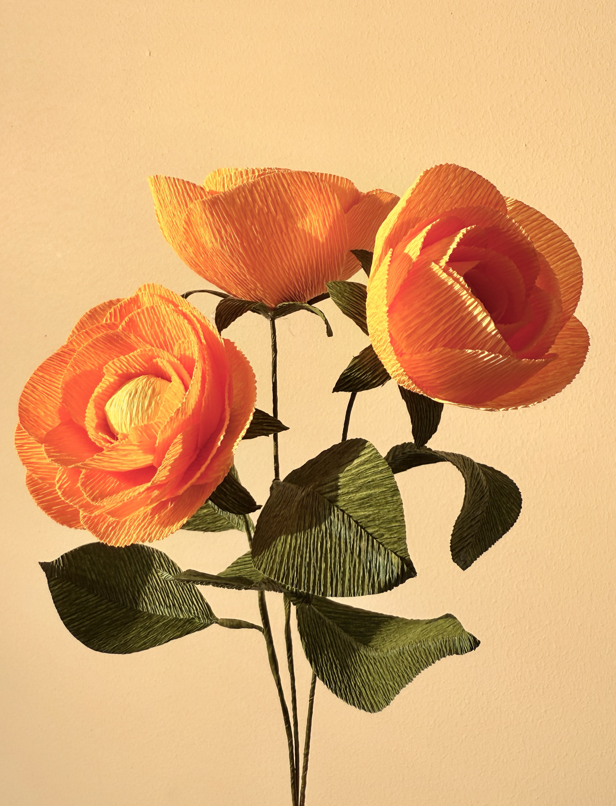 A bouquet of three orange artificial roses with green leaves set against a beige background.