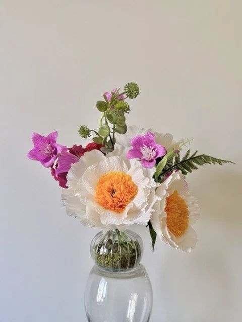 A glass vase with a colorful flower arrangement including pink, white, orange, and green blooms and foliage, set against a plain light background.