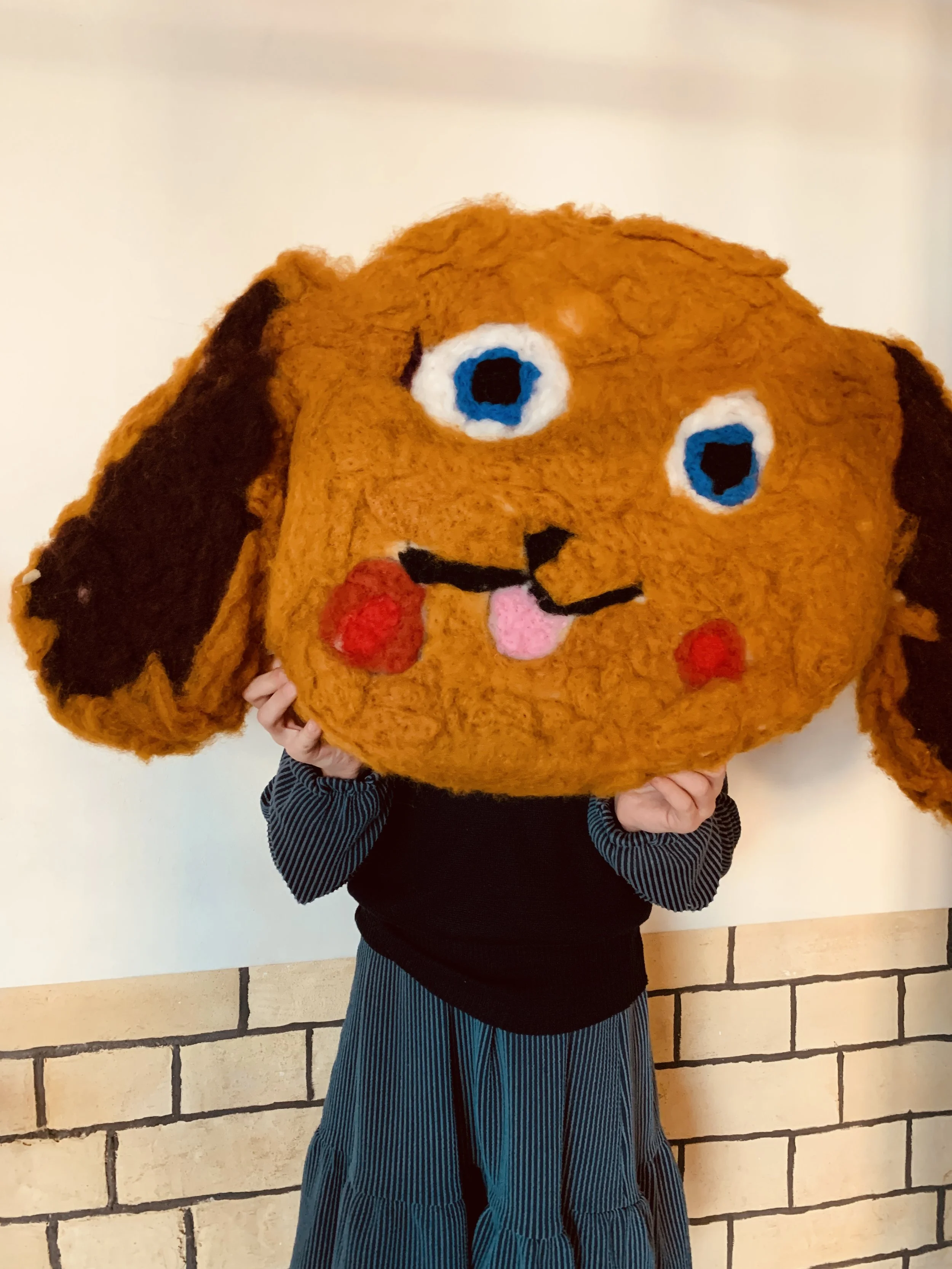 Child holding a large wool felted dog with big blue eyes, a pink tongue, and a black nose, standing in front of a beige brick wall.