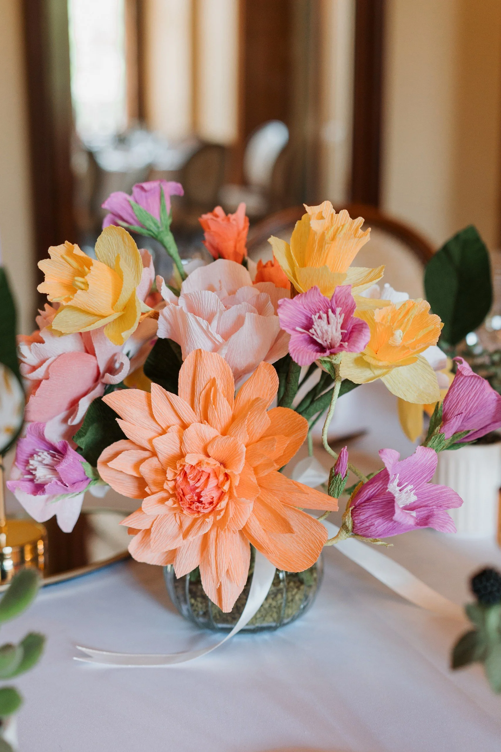 Colorful paper flower arrangement in a clear glass vase placed on a table with a white tablecloth, blurred background with wooden furniture and natural light.