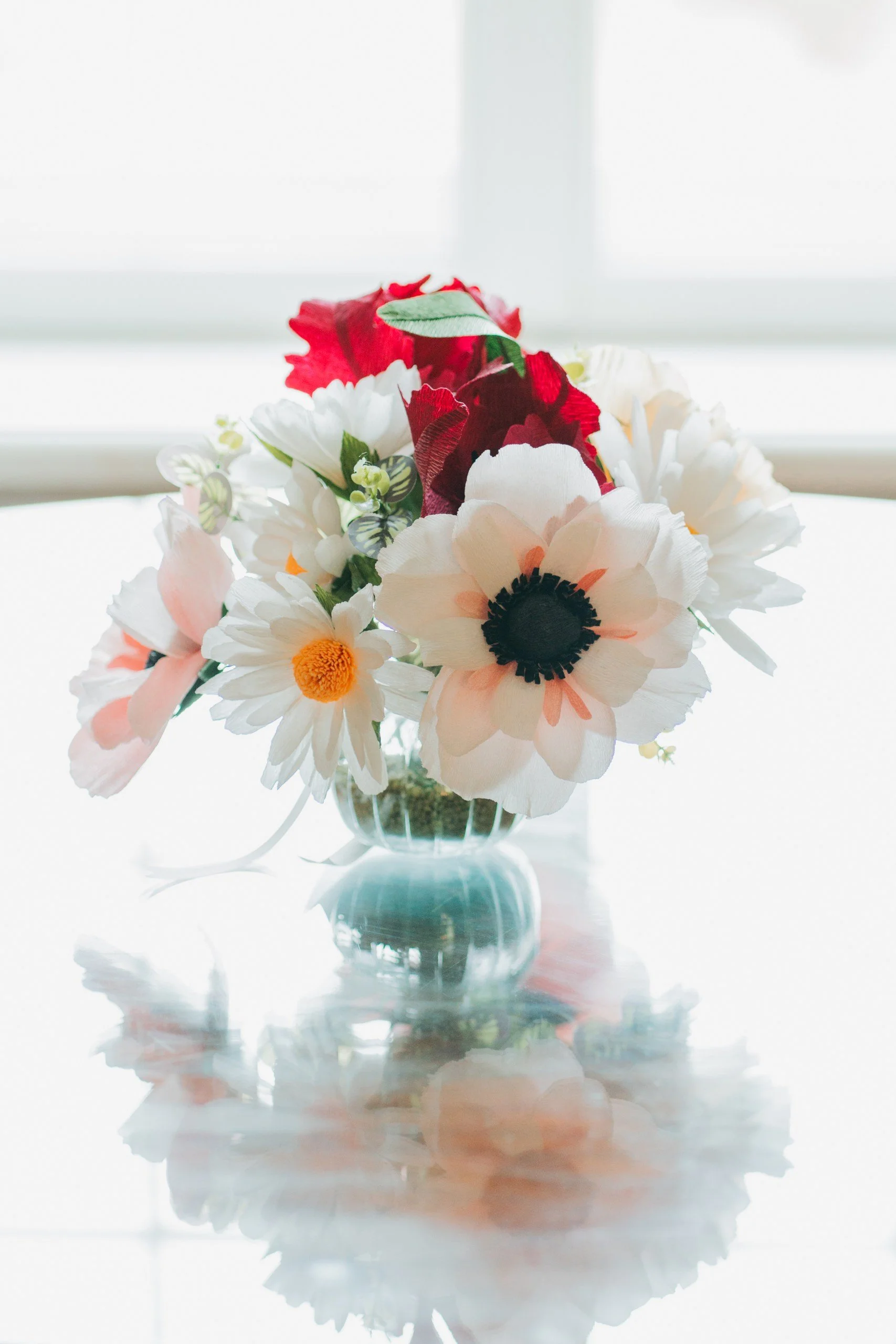 A bouquet of artificial flowers in a clear glass vase, with white, red, pink, and yellow blossoms, placed on a reflective surface with a blurred white and pale blue background.