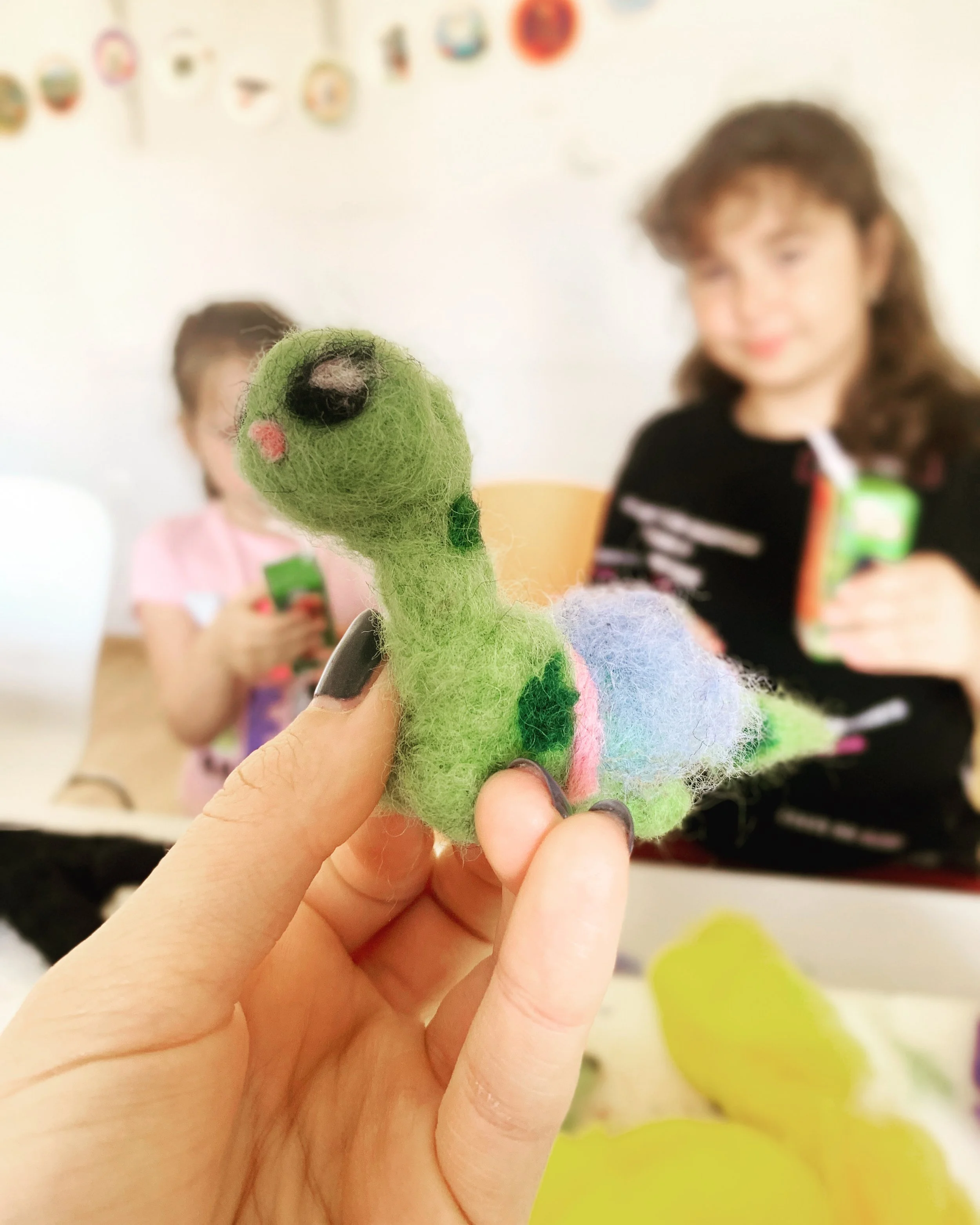 Close-up of a hand holding a green felt craft fox with multicolored felt and wool accents, in a room with children in the background.