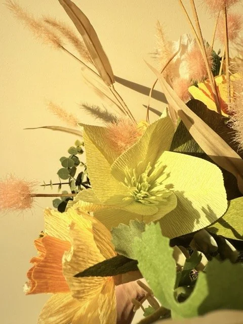 Close-up of a colorful flower arrangement with yellow, orange, and pink flowers and various green foliage, including leaves and branches.