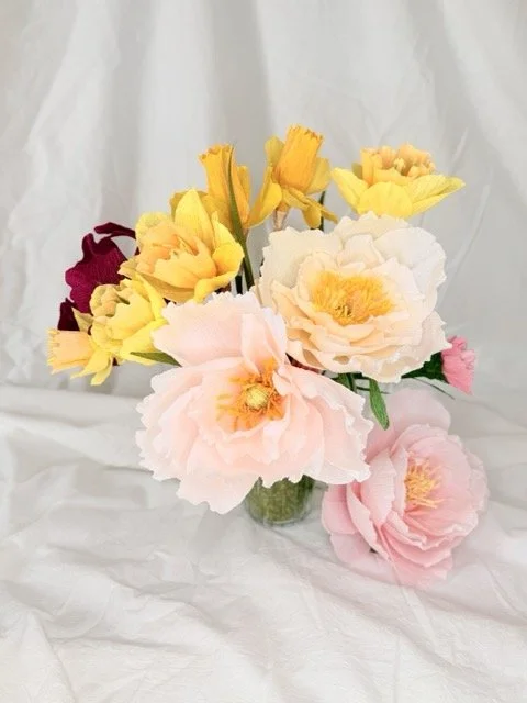 A bouquet of colorful artificial flowers in a small glass vase on a white cloth background.