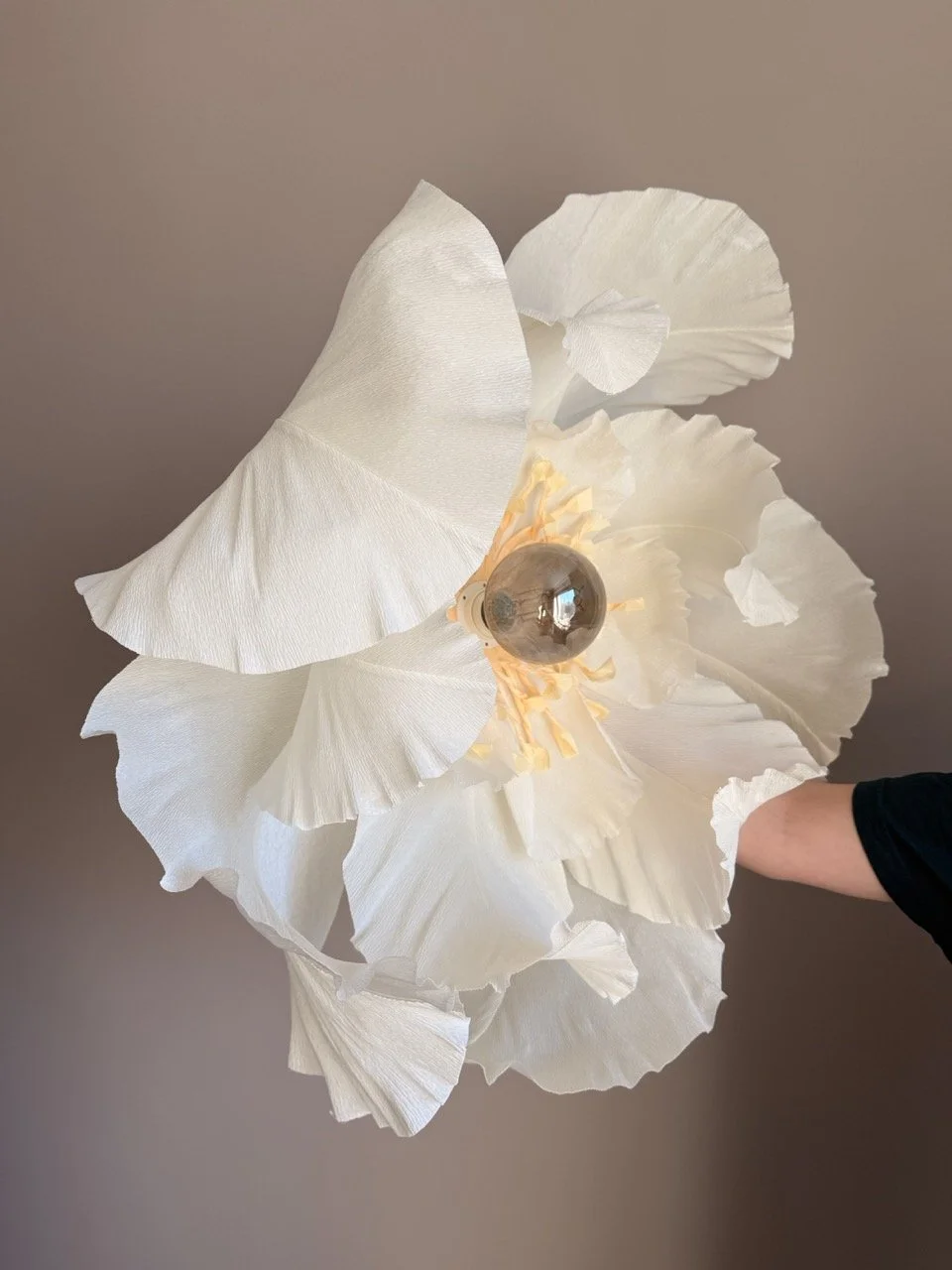 A person holding a modern, floral-inspired pendant lamp with white, ruffled paper petals and a visible light bulb in the center, against a plain beige wall.