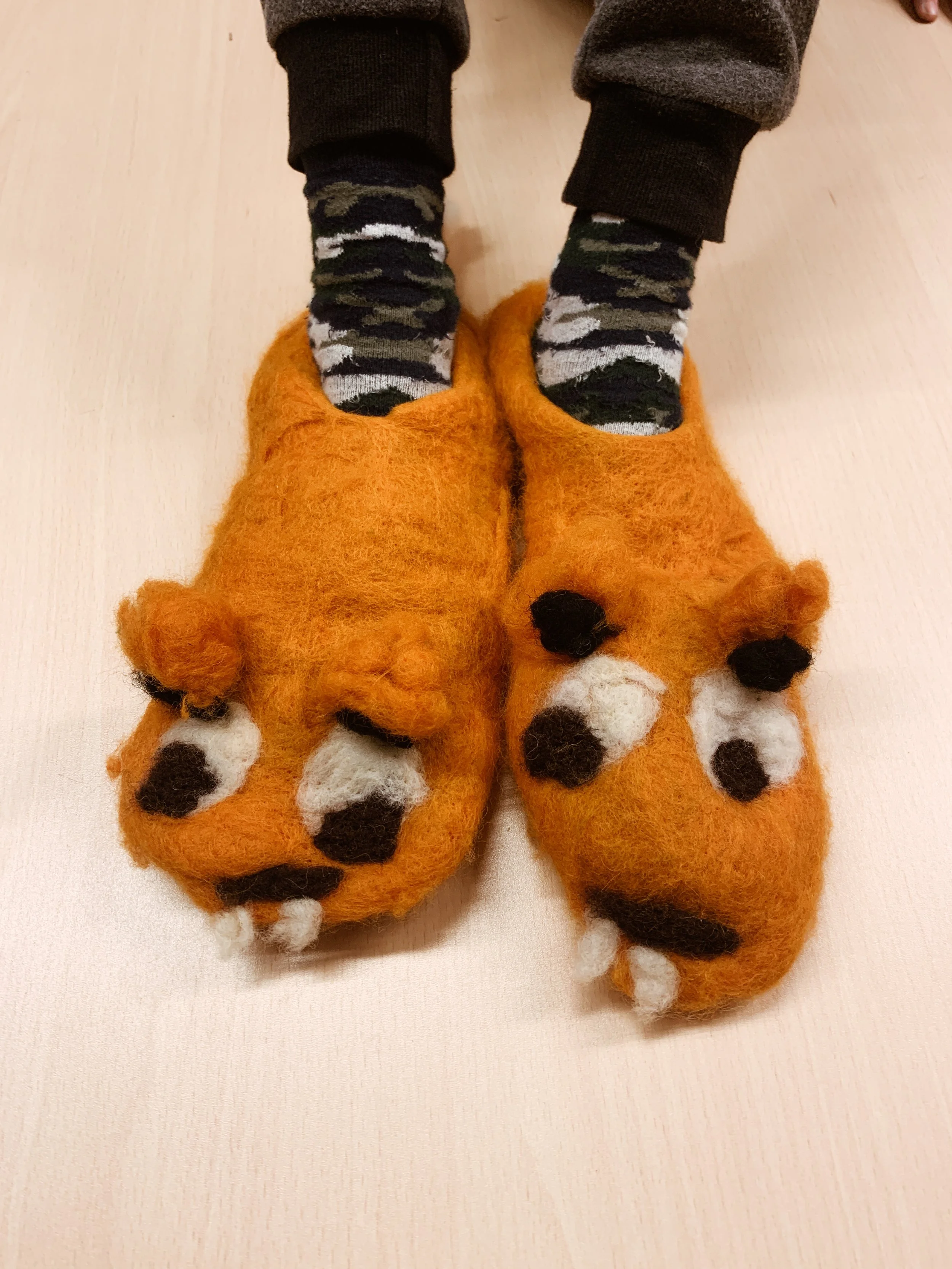 Child wearing camouflage socks and large orange fox slippers with black and white accents, on a light wooden floor.