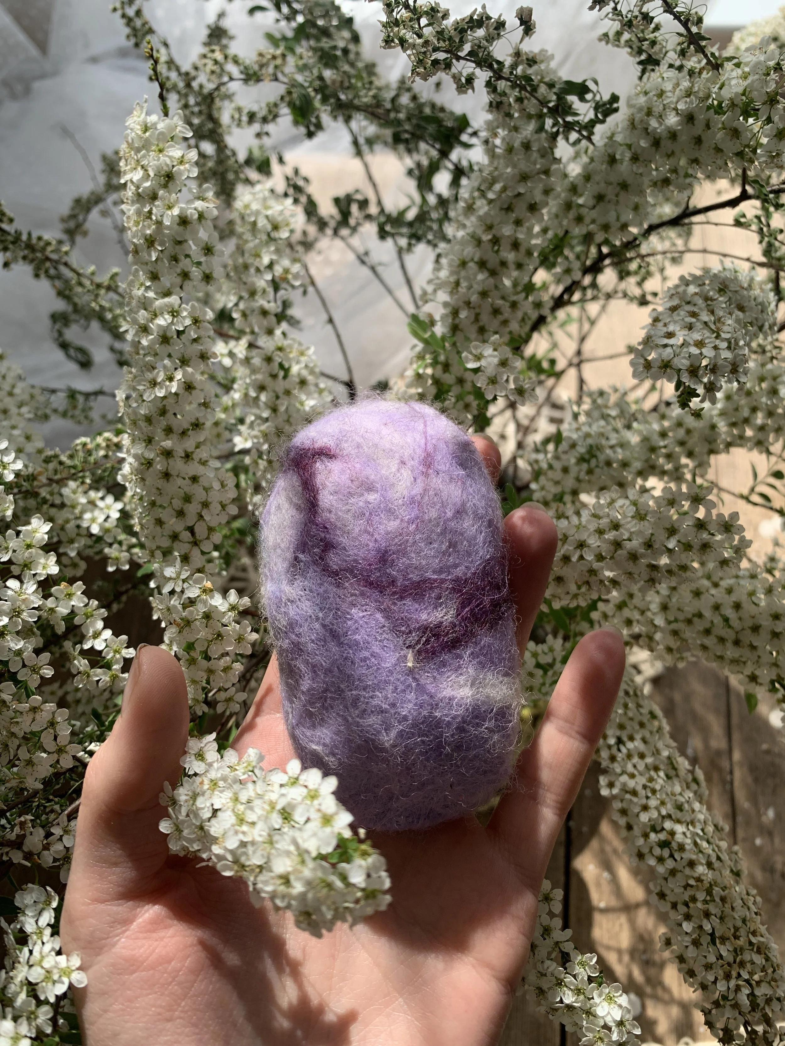 A hand holding a fuzzy, multicolored felted soap with purple, pink, and white shades, in front of a bush with small white flowers.