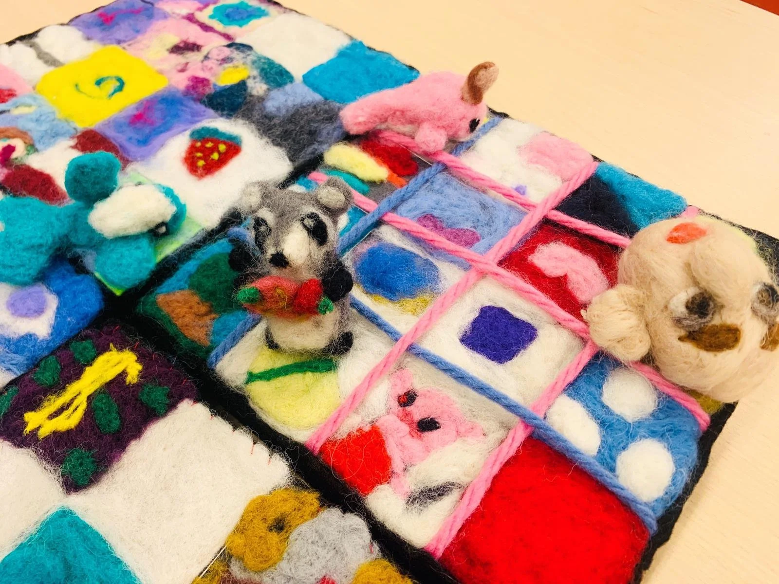 Colorful felted art pieces and small animal figurines arranged on a grid with pink yarn borders.