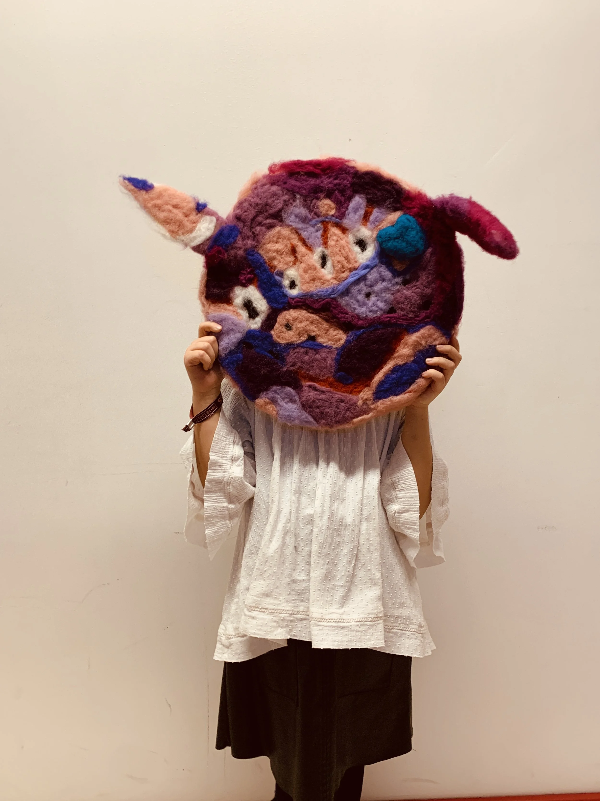 Person holding a colorful handmade felted wool wall hanging featuring a cartoon-style fox face with a unicorn horn, set against a plain beige wall.