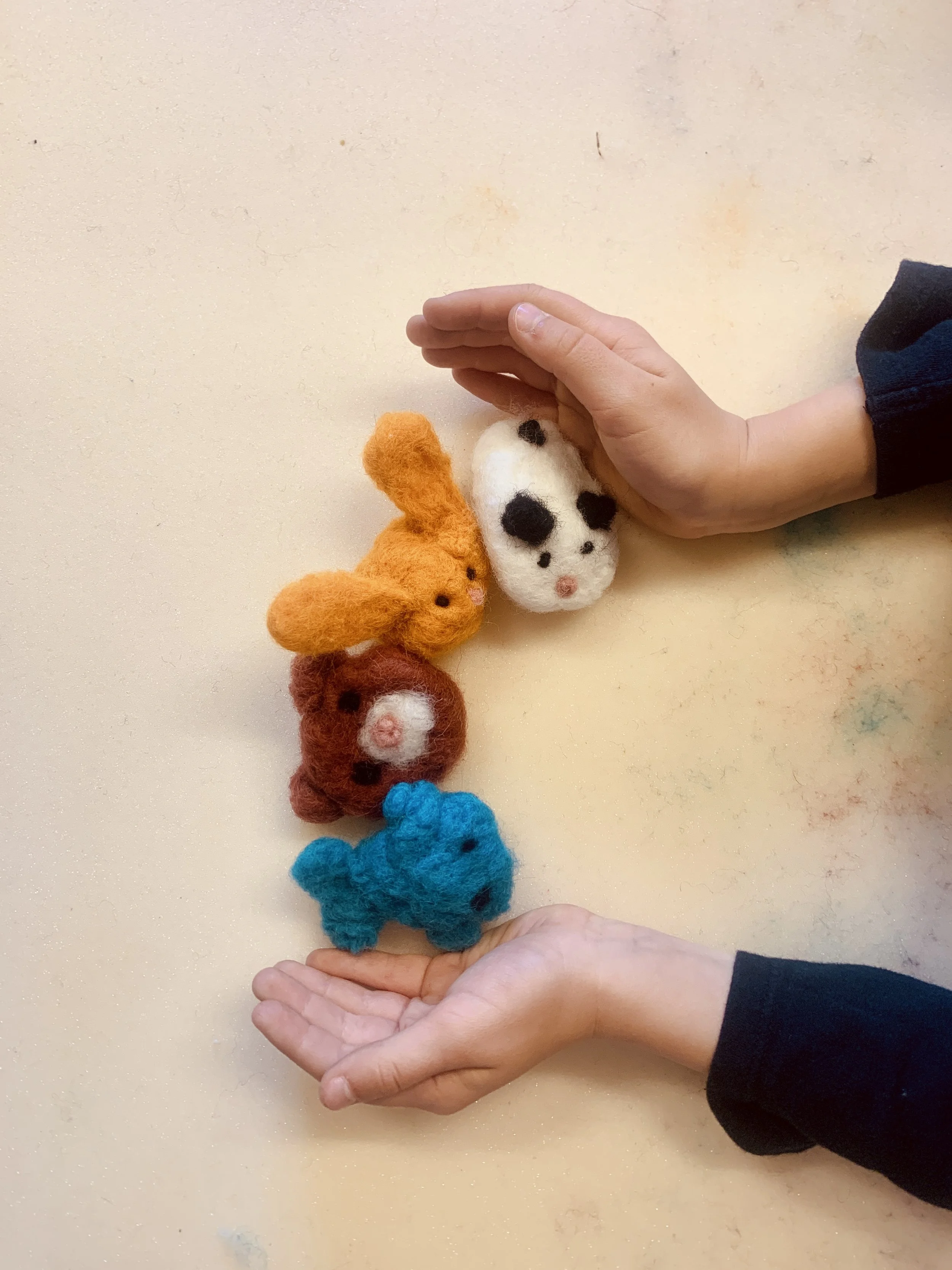 A person's hand is placed above a row of colorful felt animal figures, arranged in a line, on a light-colored surface.