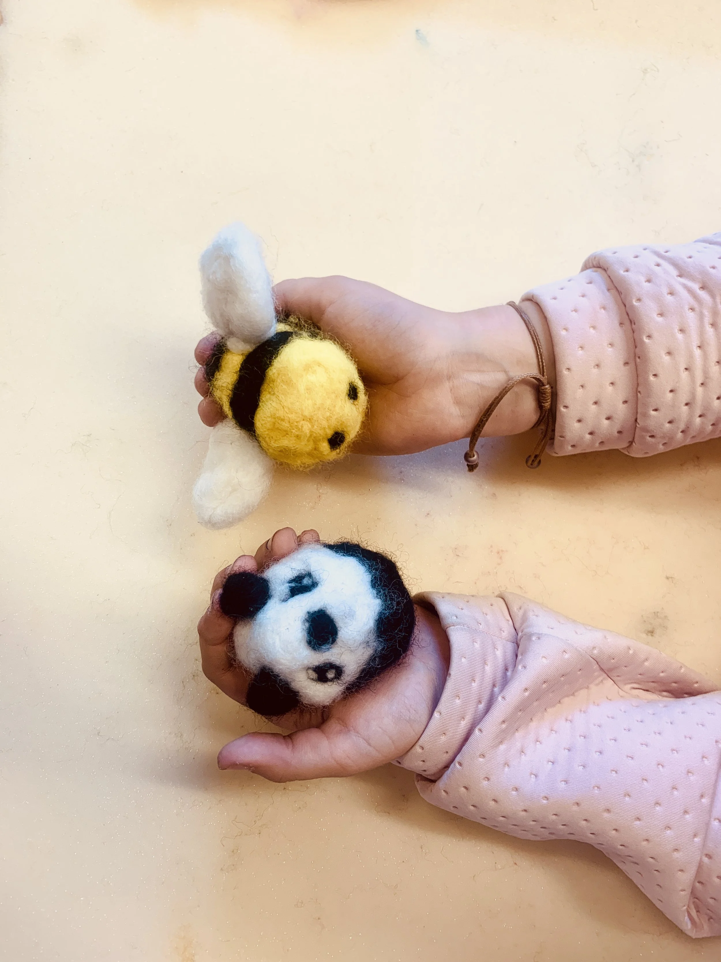 Two children's hands holding felted animal toys, including a panda and a bee, on a light-colored surface. The children are wearing pink, dotted long-sleeve shirts.