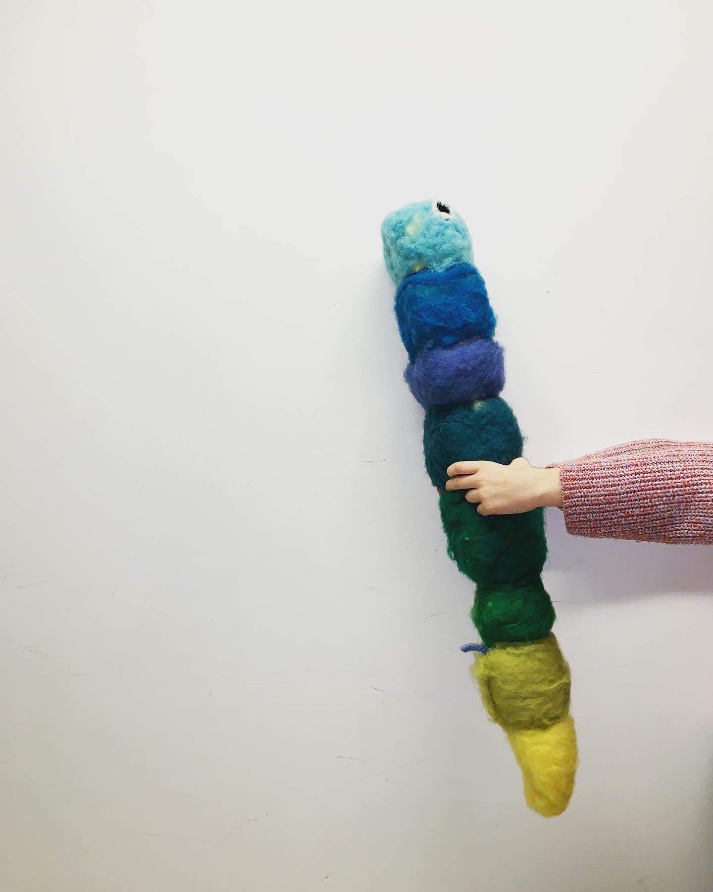 A person's arm wearing a pink sweater, holding a rainbow-colored felted wool caterpillar that is vertically arranged in rainbow order from green to yellow.