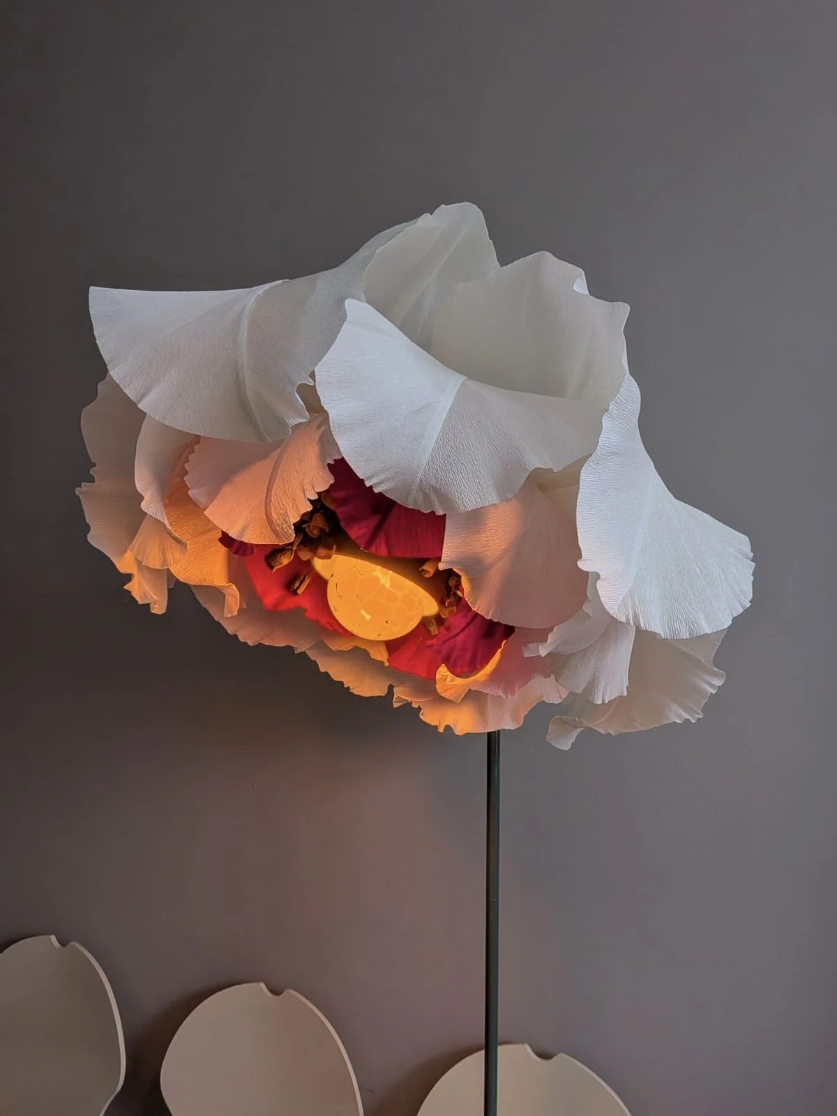 Decorative lamp designed to look like a flower with white, pink, red, and yellow petals, illuminated from within, on a stand against a plain gray wall.