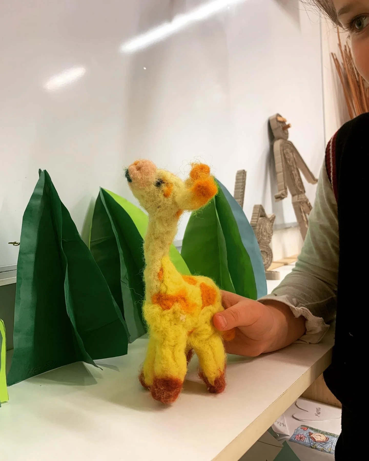 A person holding a felted giraffe toy in front of paper trees and leaf props. The background shows a plain white wall and wooden crafts.