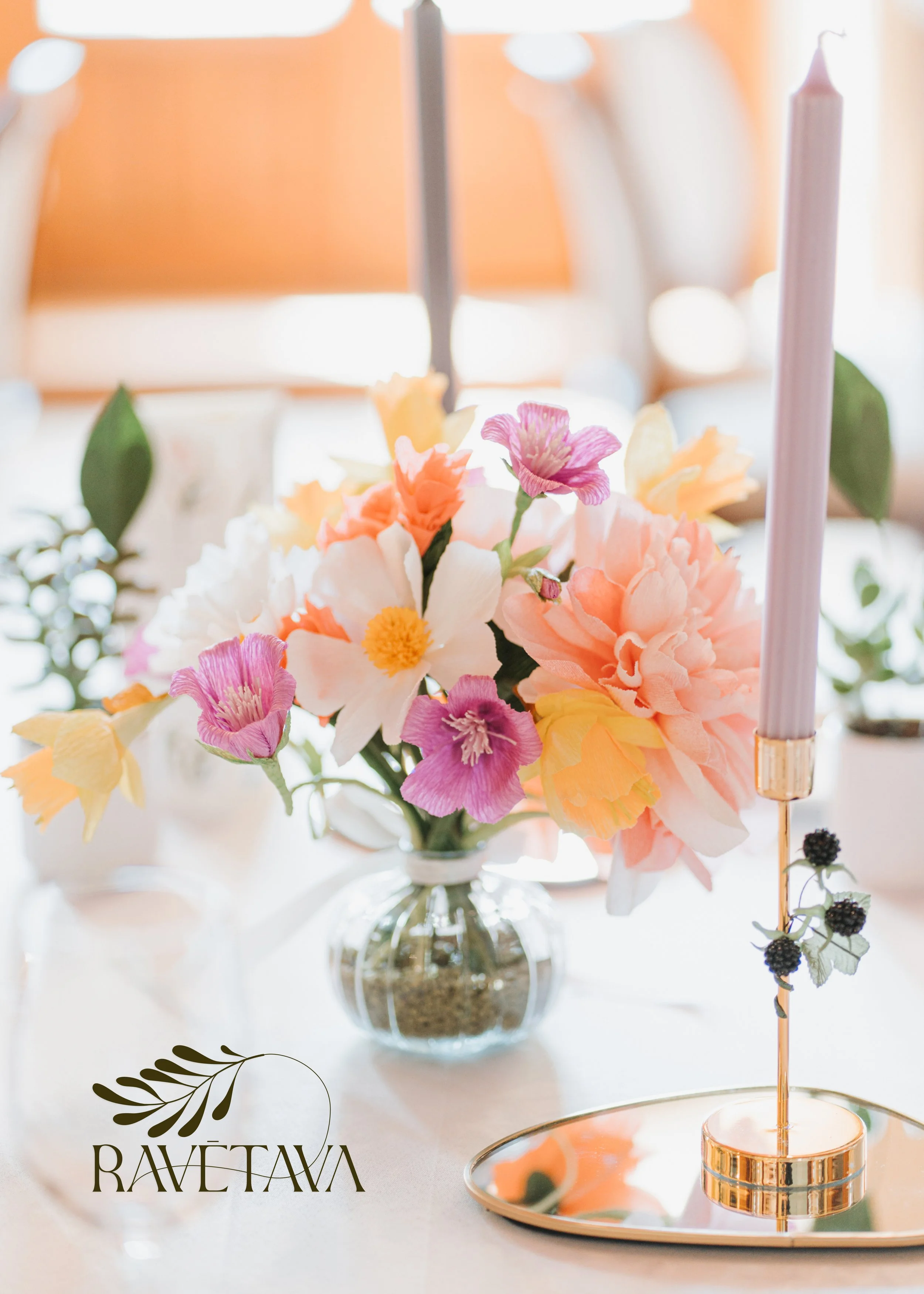 Elegant floral centerpiece with pink, peach, and white flowers in a glass vase, alongside a white candle in a gold holder placed on a silver tray. Part of a wedding table set-up, with a blurred background and a decorative logo reading 'RAVETAVA'.