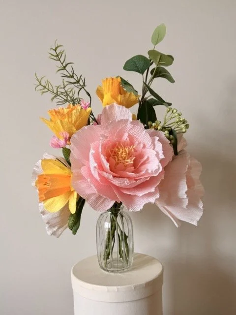 A glass vase with a bouquet of colorful paper flowers, including pink, yellow, and orange flowers, along with green leaves and foliage, placed on a white circular stand against a plain background.