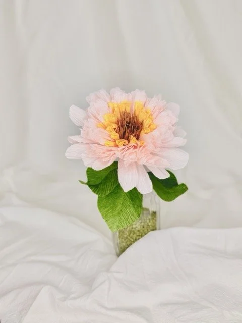A pink artificial flower with yellow and brown center in a small glass vase, placed on a white fabric background.