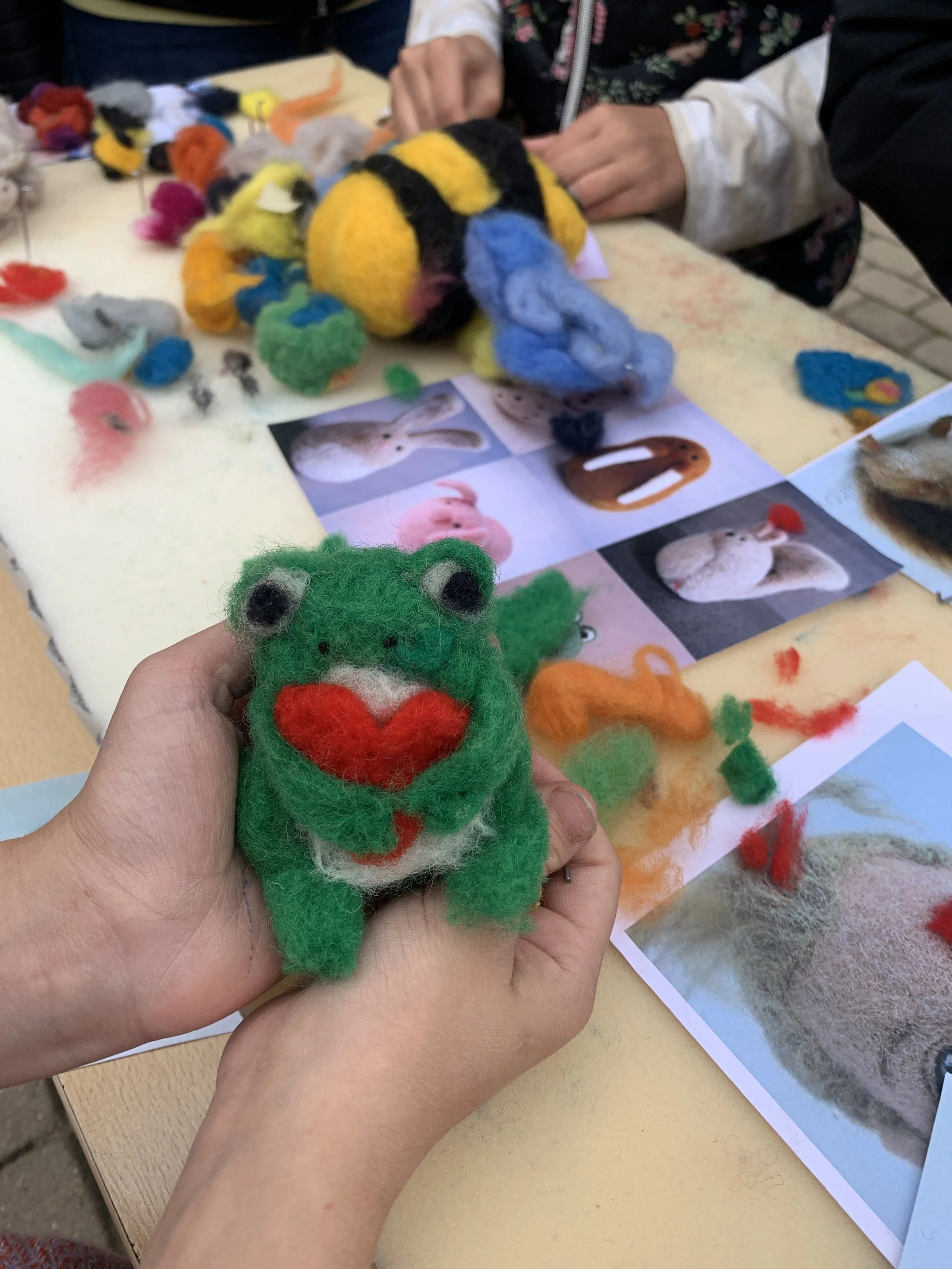 Person holding a small, colorful felted bunny toy with red heart and green body in a craft activity at a table with felted pieces and reference images of rabbits.