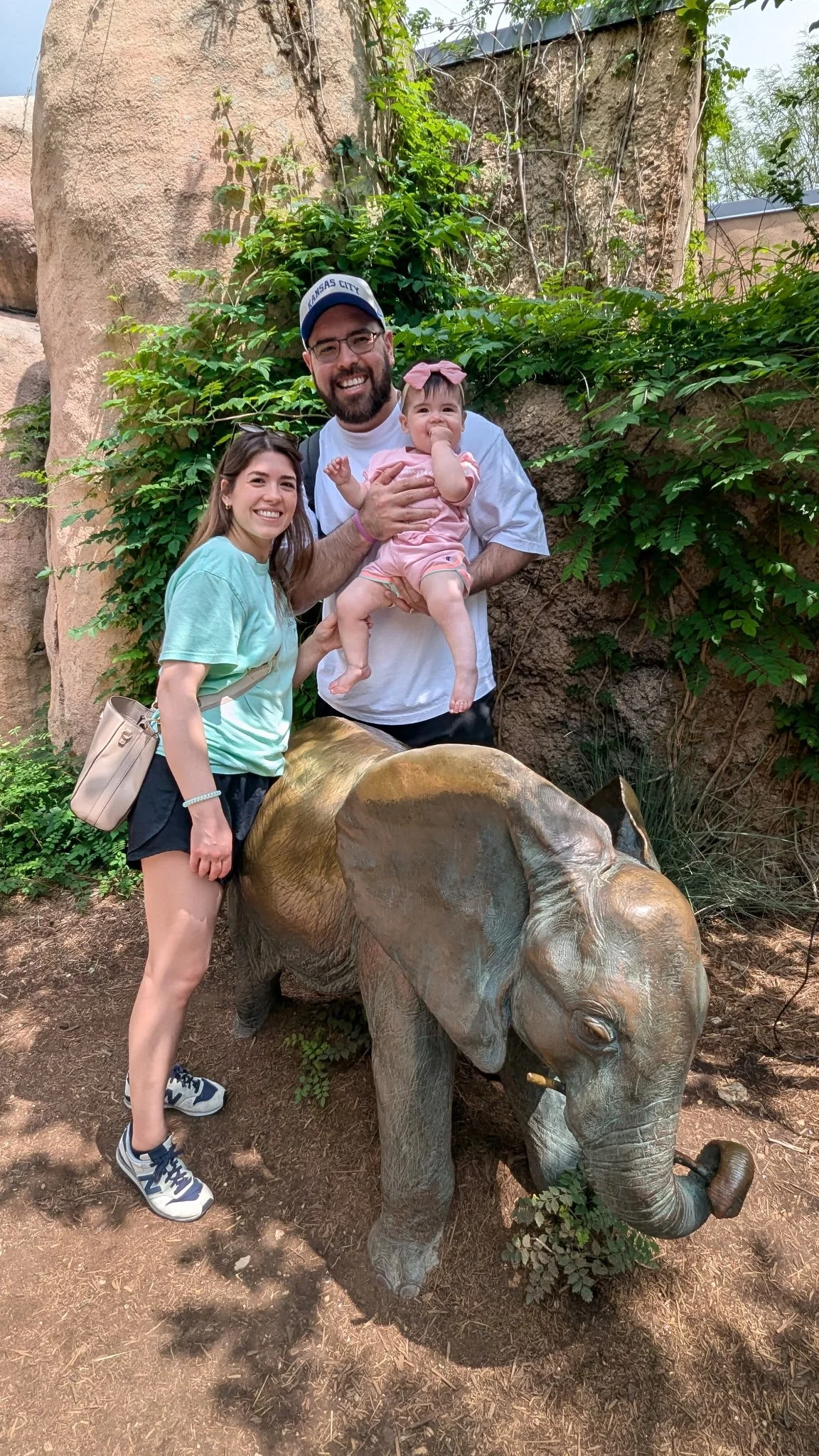 A family of three standing next to a bronze elephant statue outdoors, smiling at the camera. The father holds a young girl, and the mother stands beside them, on a dirt ground with greenery and a rock wall in the background.