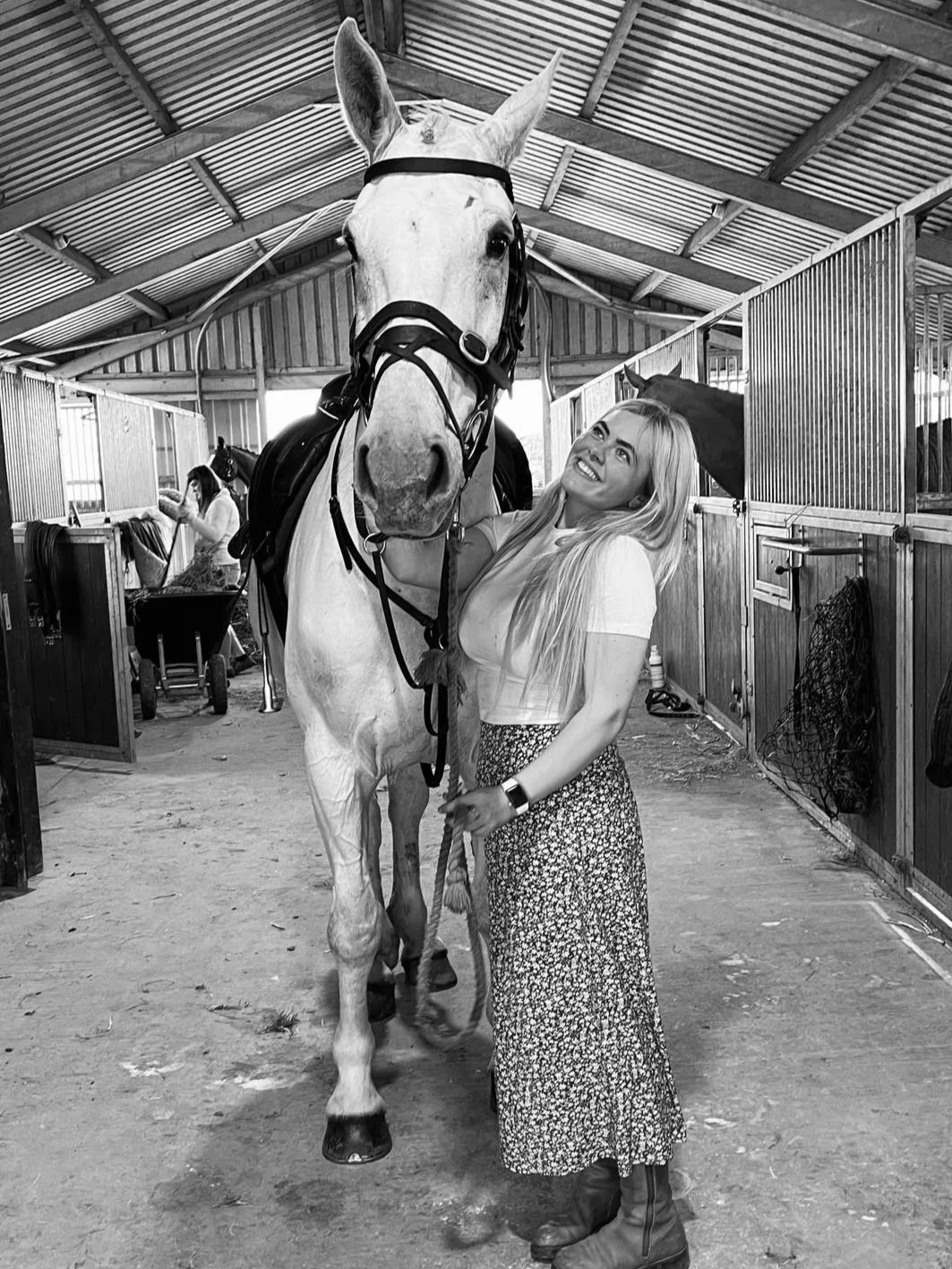 A woman with long blonde hair smiling and standing next to a large white horse inside a stable, holding its bridle.
