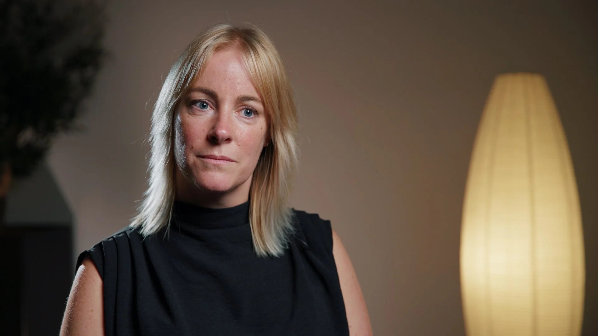 A blonde woman with blue eyes in a black sleeveless top sitting indoors, with a beige lamp in the background.