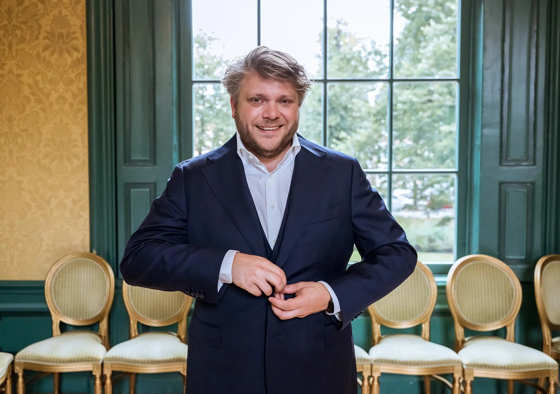 Iman de Vries-Maarse in a dark blue suit and white shirt standing in a room with large windows with green trim and yellow walls, smiling and adjusting his cufflinks.