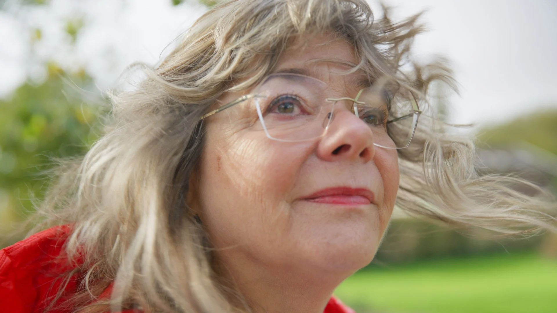 Close-up of a woman with curly blonde hair wearing glasses and a red jacket outdoors, with a blurred background of greenery.