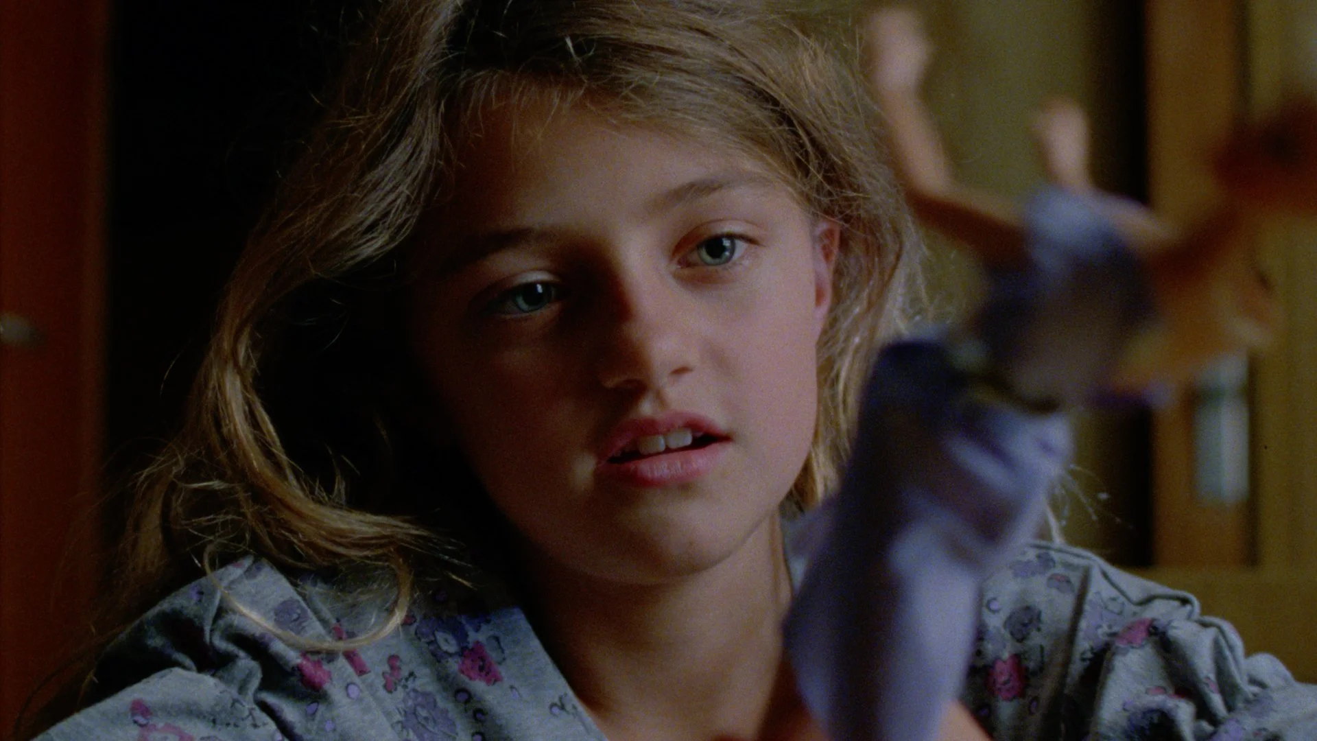Close-up of a young girl with light brown hair and blue eyes, looking thoughtfully while touching or pointing at something off-camera.
