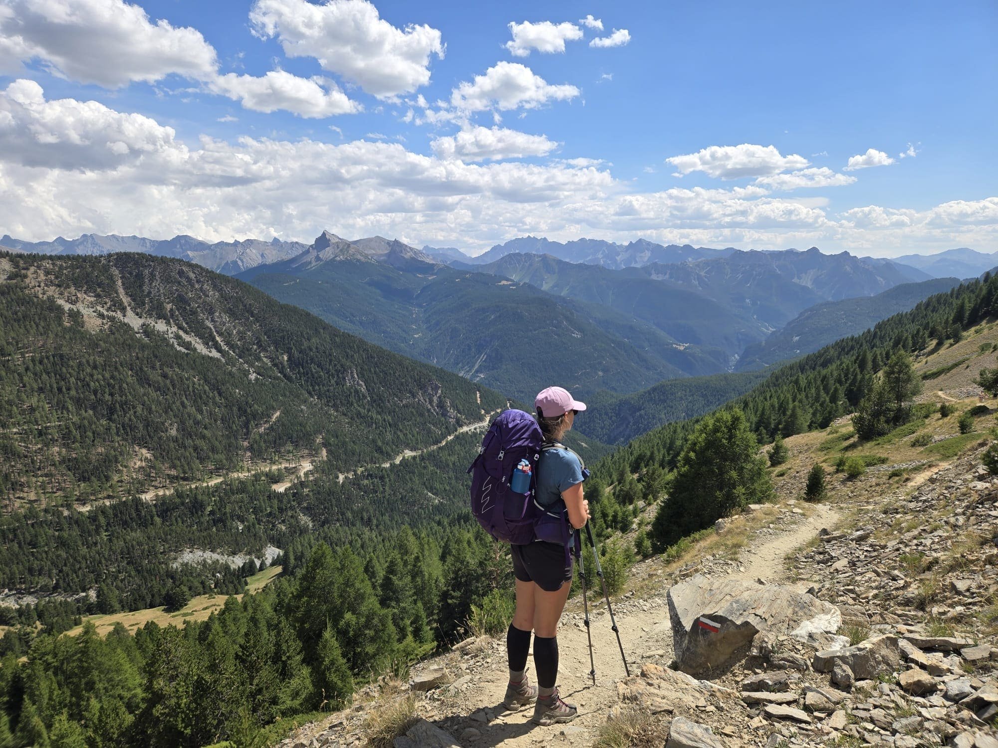Une personne portant un chapeau rose, une veste bleue, un sac à dos violet, tenant des bâtons de marche, regarde la montagne dans un paysage de montagnes escarpées et forêt sous un ciel partiellement nuageux.