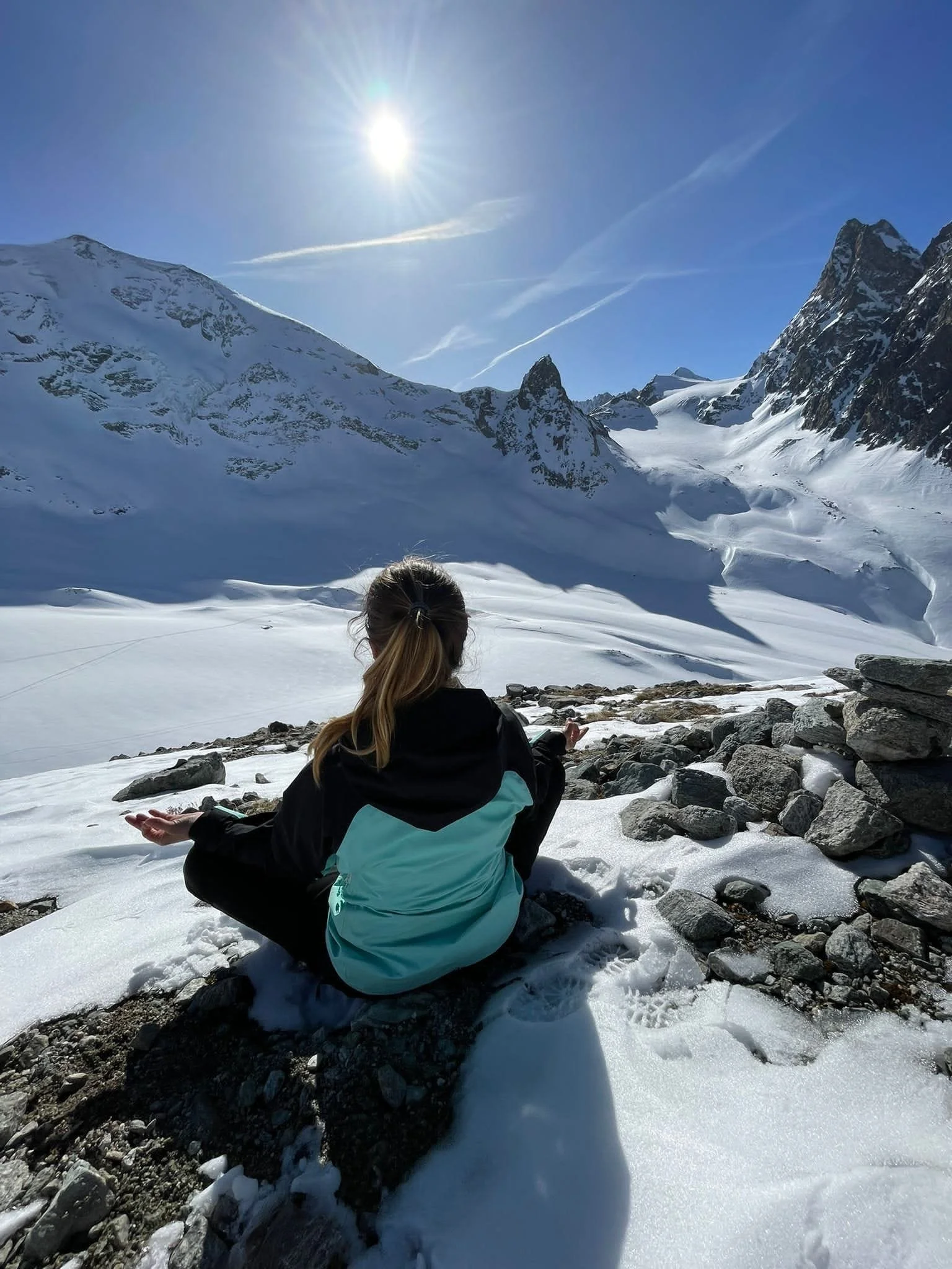 Une personne assise en méditation ou contemplation dans un paysage de montagne enneigée avec un ciel bleu clair et le soleil brillant haut dans le ciel.