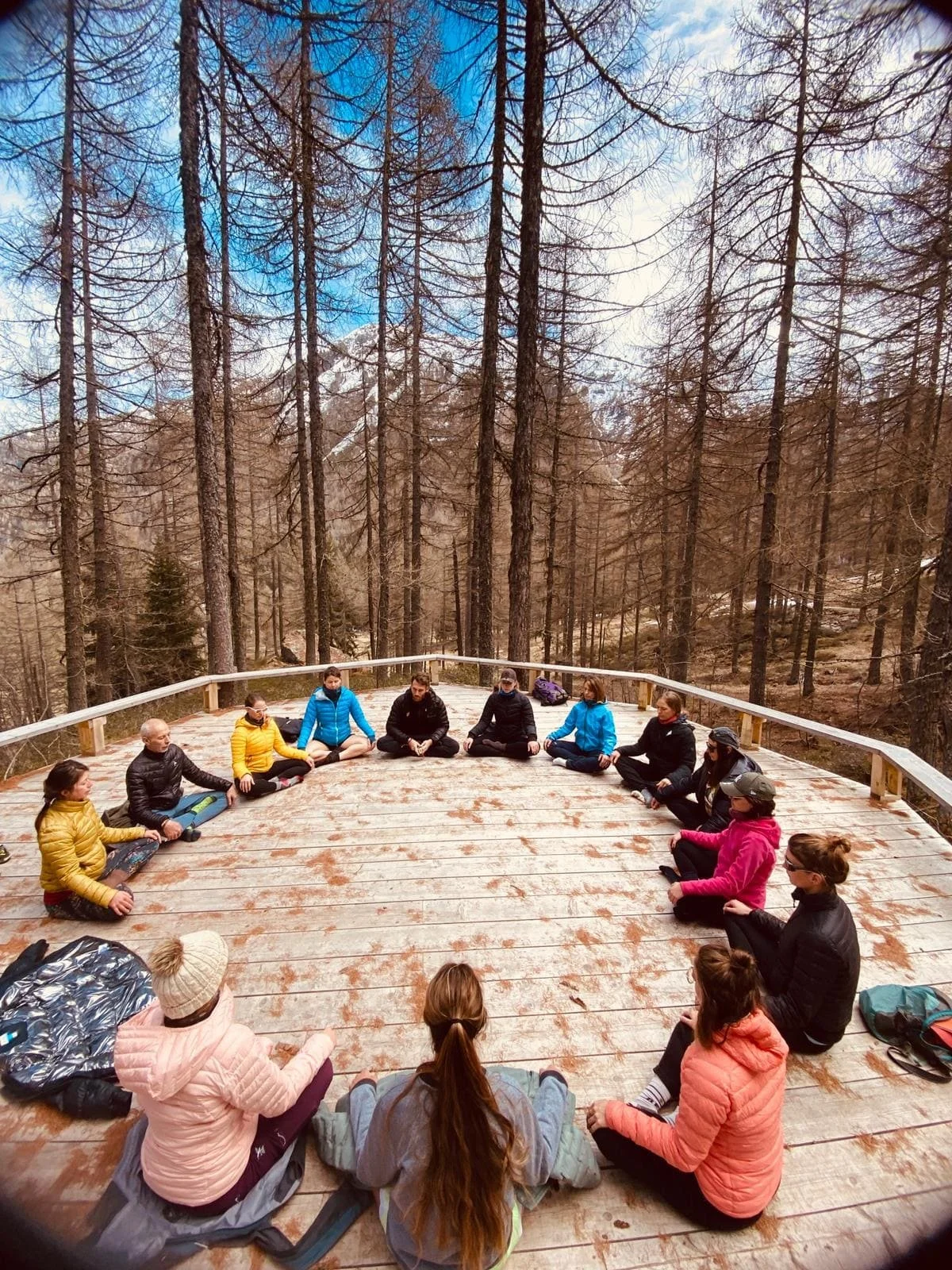 Groupe de personnes assises en cercle sur une plateforme en bois entourée d'arbres dans une forêt.