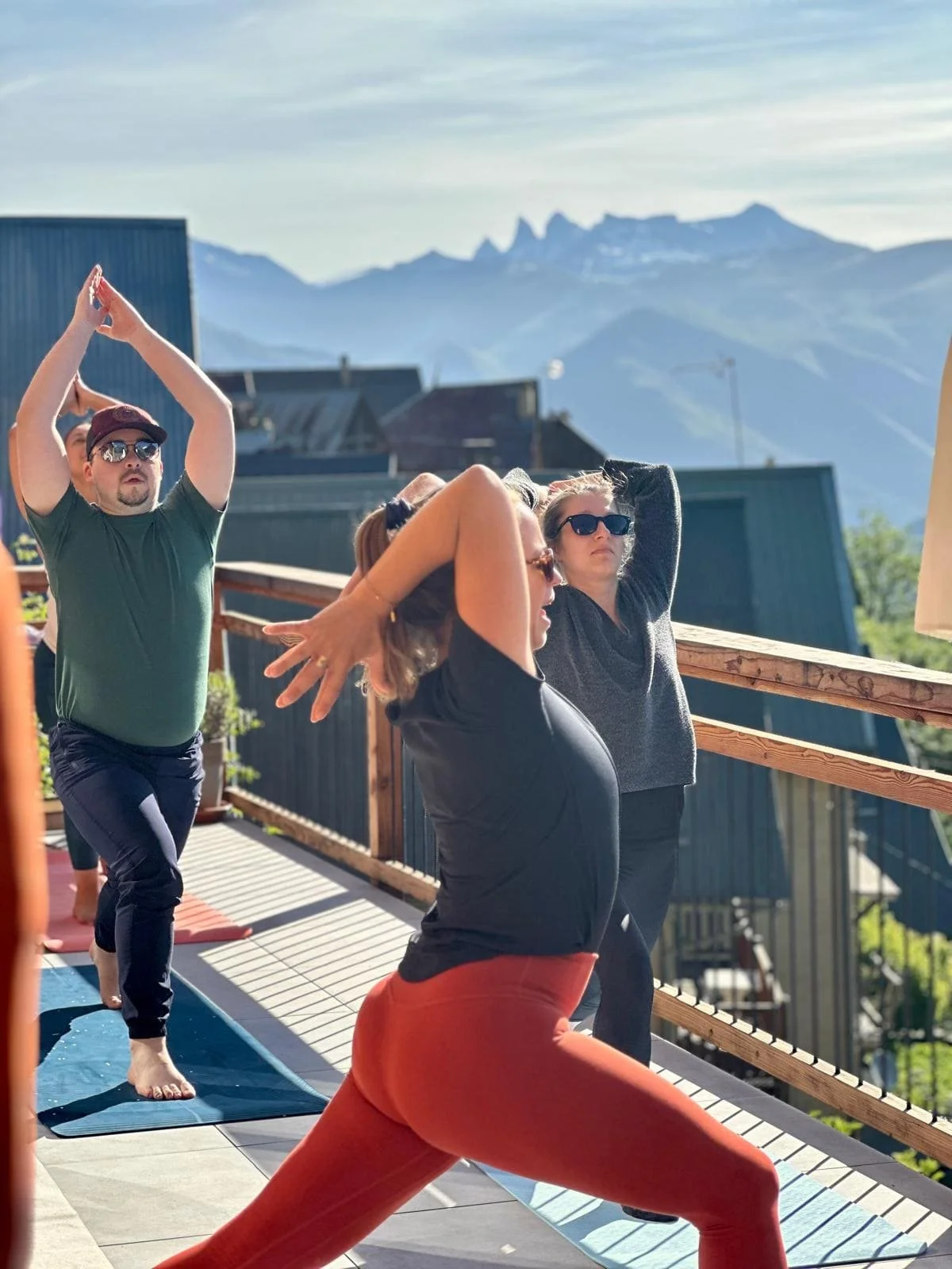 Groupe de personnes pratiquant le yoga sur une terrasse extérieure, avec une vue sur des montagnes en arrière-plan.