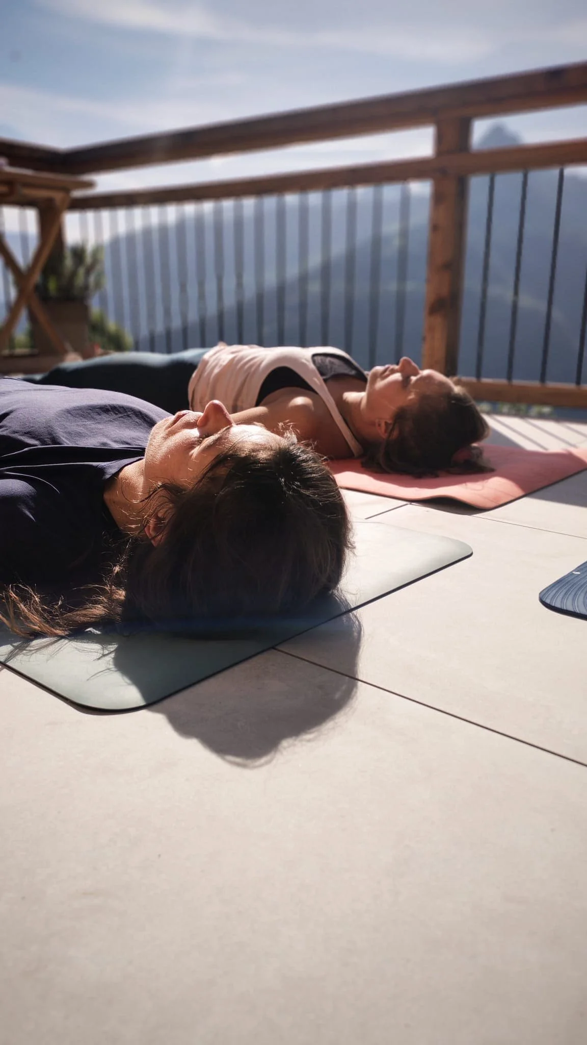 Deux femmes font du yoga allongées sur des tapis, en plein air sur une terrasse en bois avec une vue sur des montagnes.