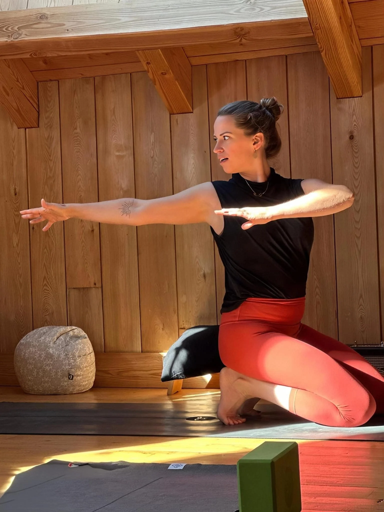 Laura, professeure de yoga en position de pose de la chameau dans une pièce en bois, avec un coussin, un gros rocher et des blocs de yoga