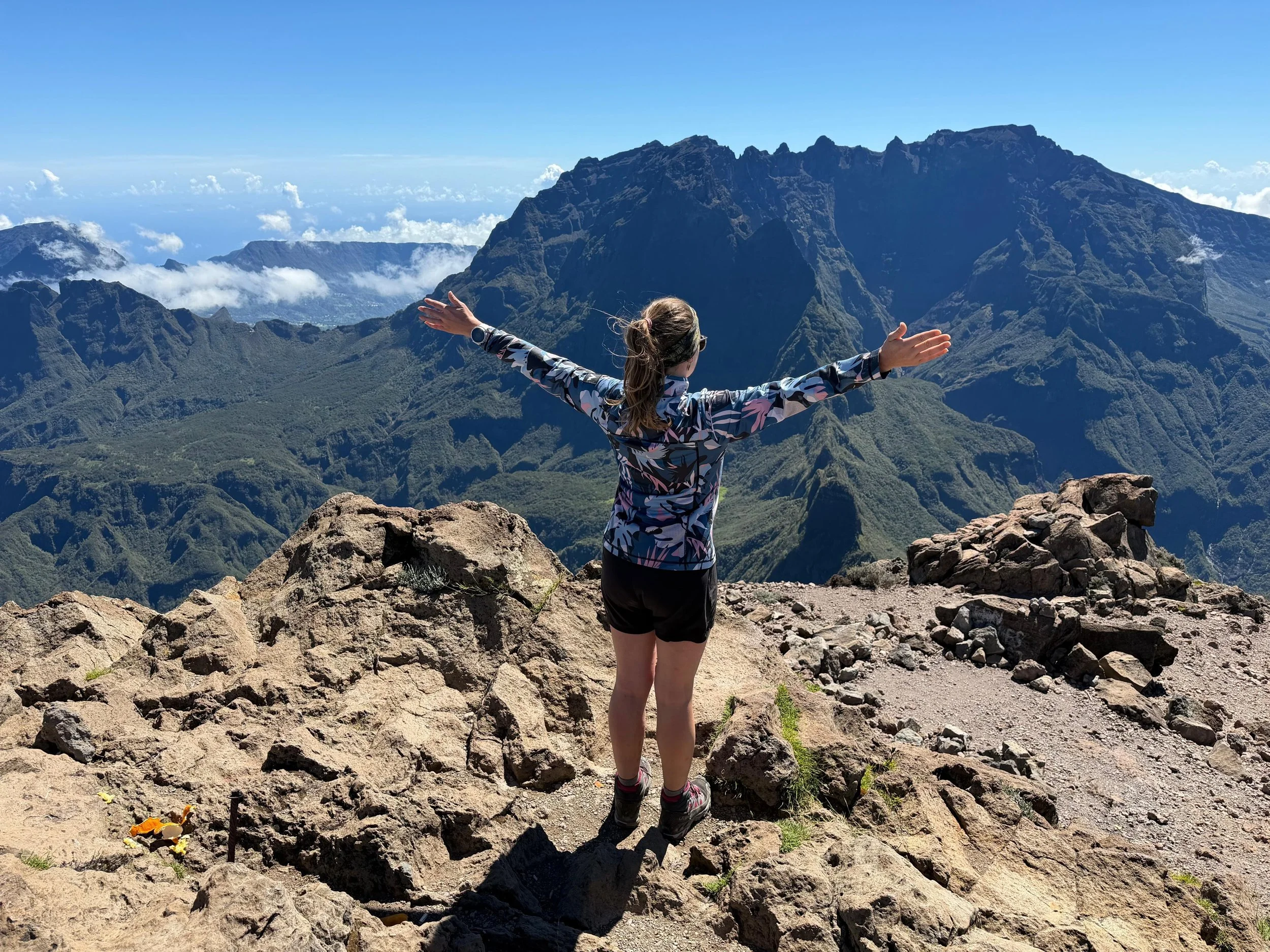 Une personne debout sur un rocher, bras levés, regardant un paysage de montagnes majestueuses sous un ciel bleu clair, avec des nuages en arrière-plan.