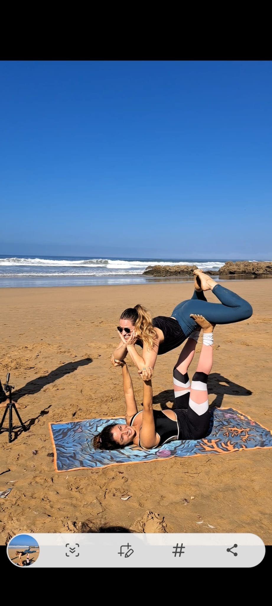 Deux femmes font du yoga sur la plage, une d'elles est allongée sur un tapis et a soulevé l'autre au-dessus d'elle, qui sourit, en position de yoga, avec la mer en arrière-plan.