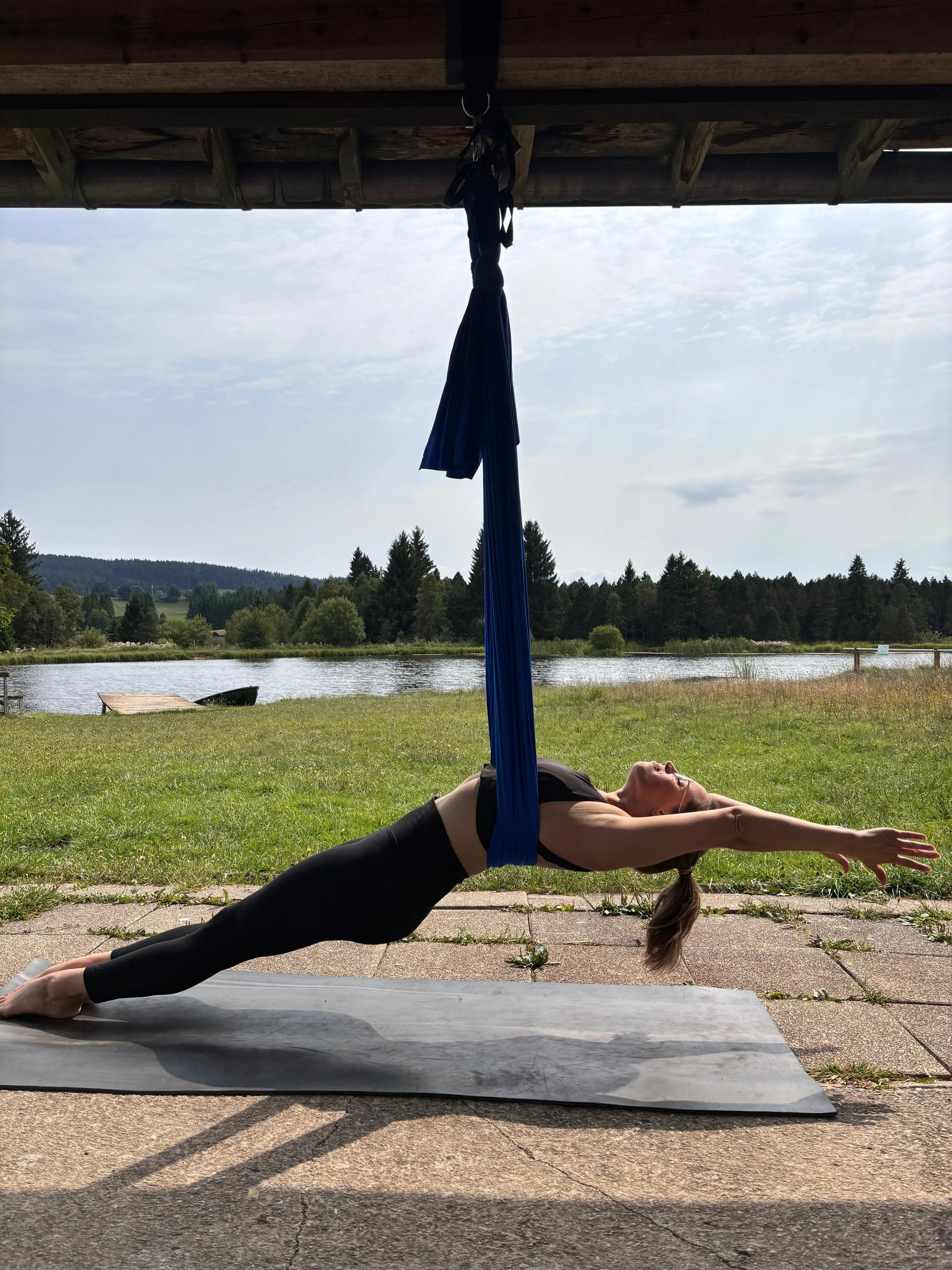 Une femme pratique le yoga en position de planche, allongée sur un tapis, sous une structure en bois avec une vue sur la nature et un lac en arrière-plan.