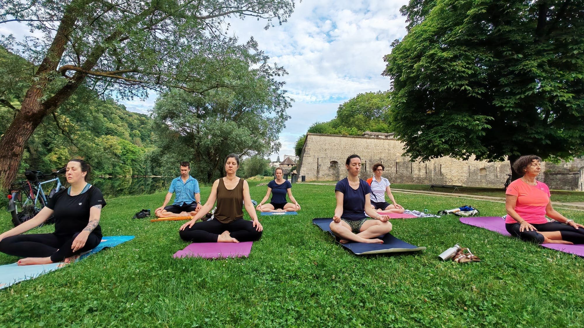 Groupe de personnes pratiquant la méditation ou le yoga en plein air sur une pelouse verte, avec des arbres et un bâtiment en pierre en arrière-plan.