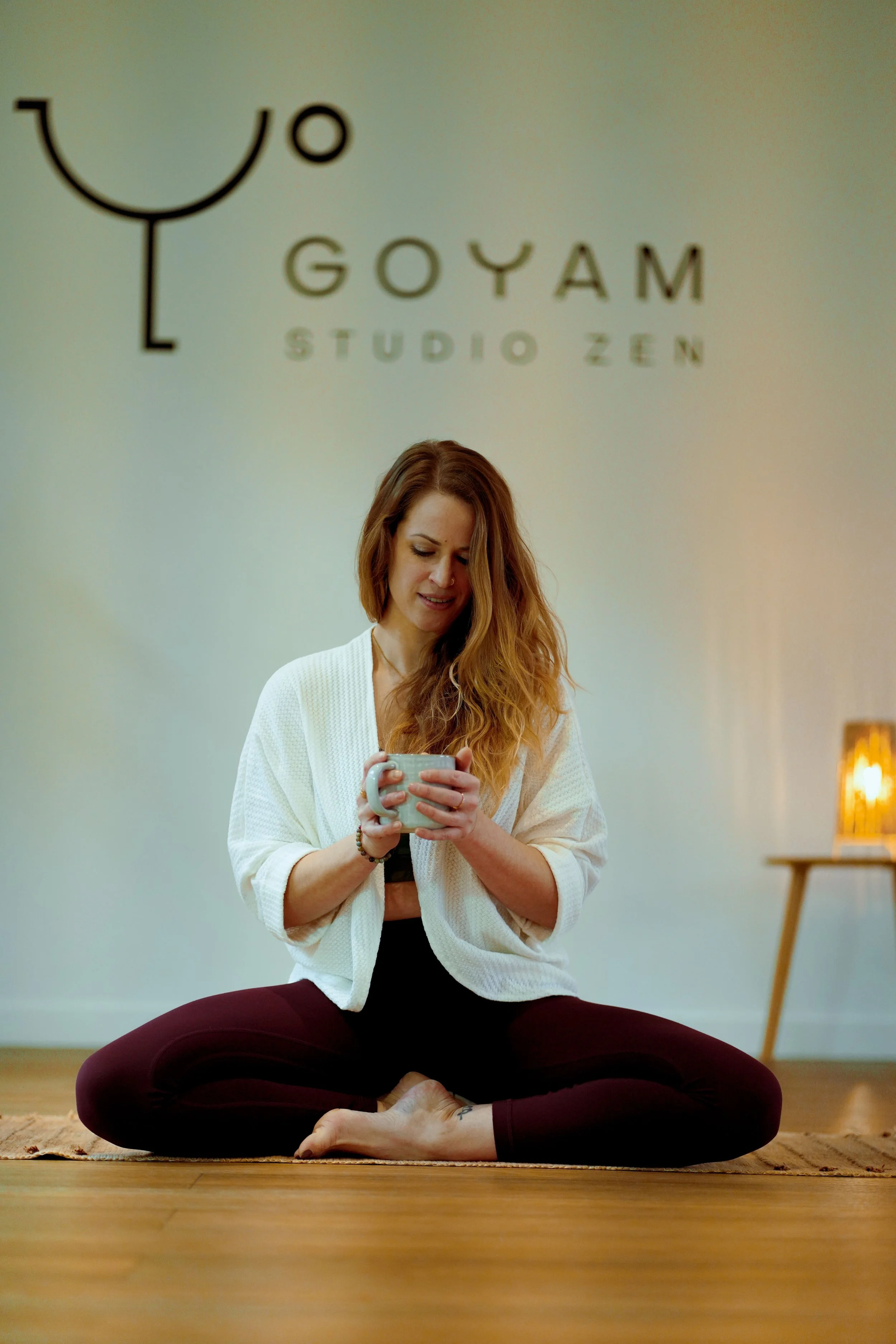 Une femme assise en position de lotus sur un tapis, tenant une tasse, en tenue décontractée, dans une pièce avec une lumière douce, une table en bois à droite, et un logo de 'Goyam Studio Zen' en arrière-plan.