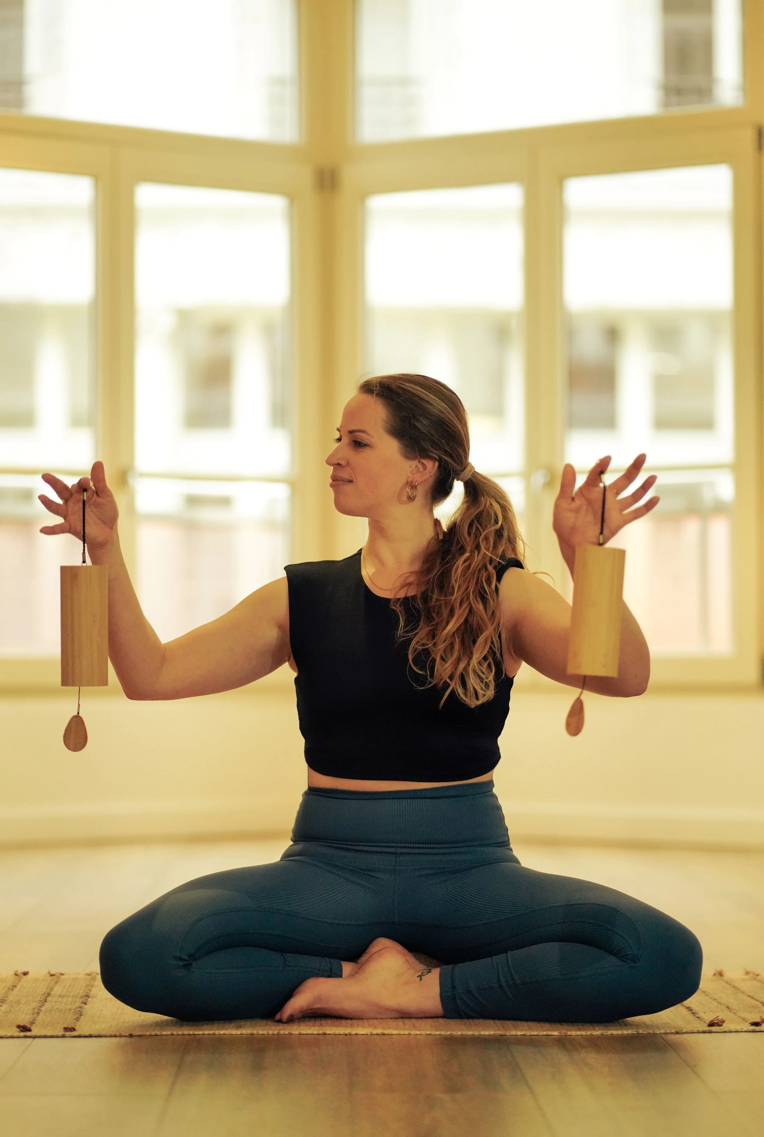 Laura donnant un cours de yoga en position de médiation en tailleur dans une pièce lumineuse avec de grandes fenêtres. Elle porte un haut noir et un pantalon de sport gris foncé, et est assise en tailleur sur un tapis.