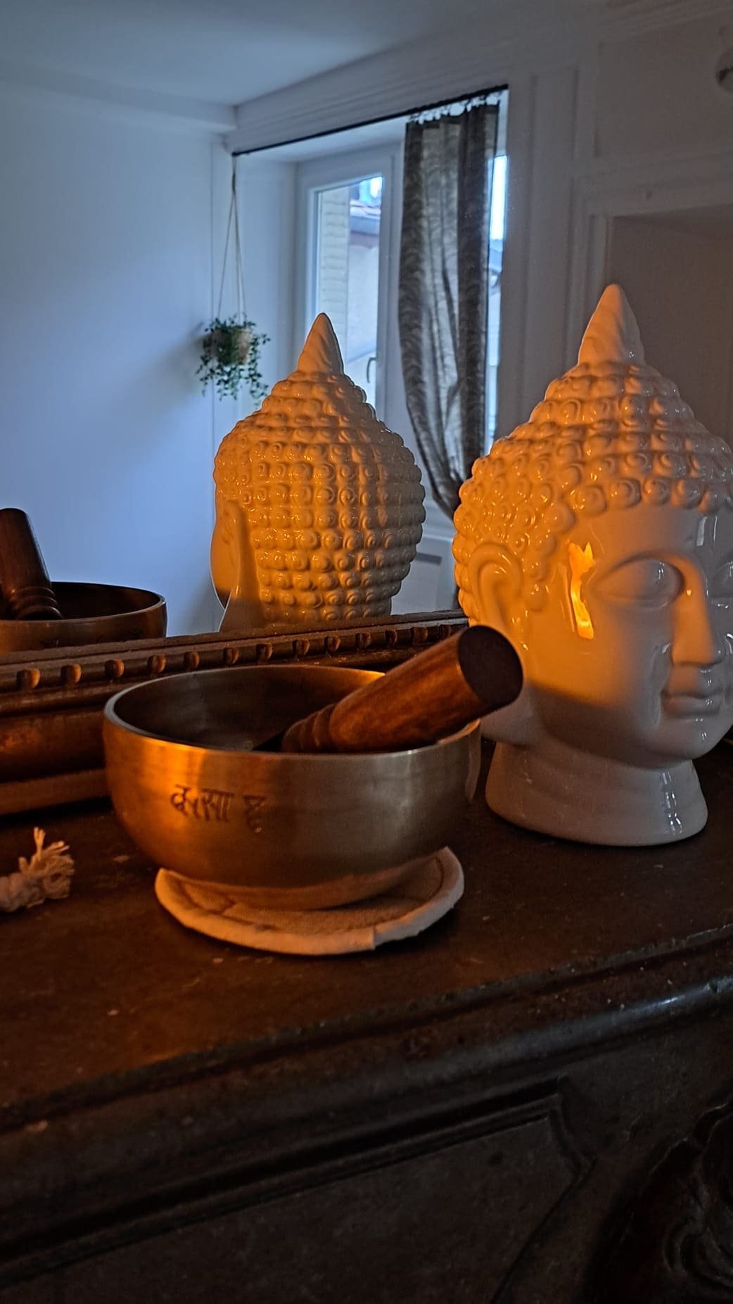 Décoration intérieure avec des lampes en forme de tête de Bouddha en céramique, un bol en métal avec un marteau en bois à l'intérieur, et un miroir reflétant une fenêtre avec une plante suspendue.