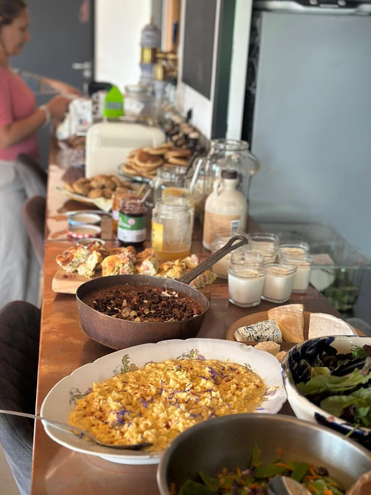 Table rempli de plats variés comme une quiche, une cocotte avec une sauce ou un ragoût, desserts, fromages, yaourts, et salades, avec des personnes en arrière-plan lors d'un repas.