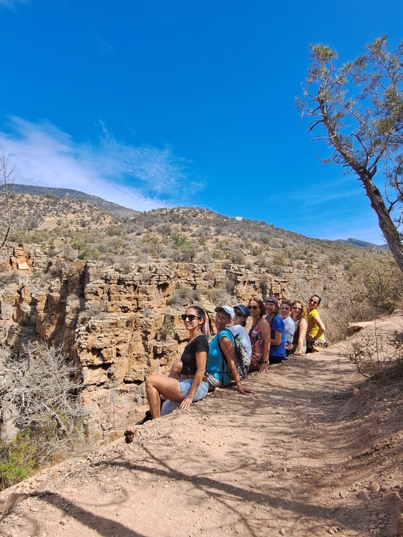 Groupe de personnes assises en rang sur un sentier rocheux en plein air, avec des montagnes arides en arrière-plan et un ciel bleu clair.