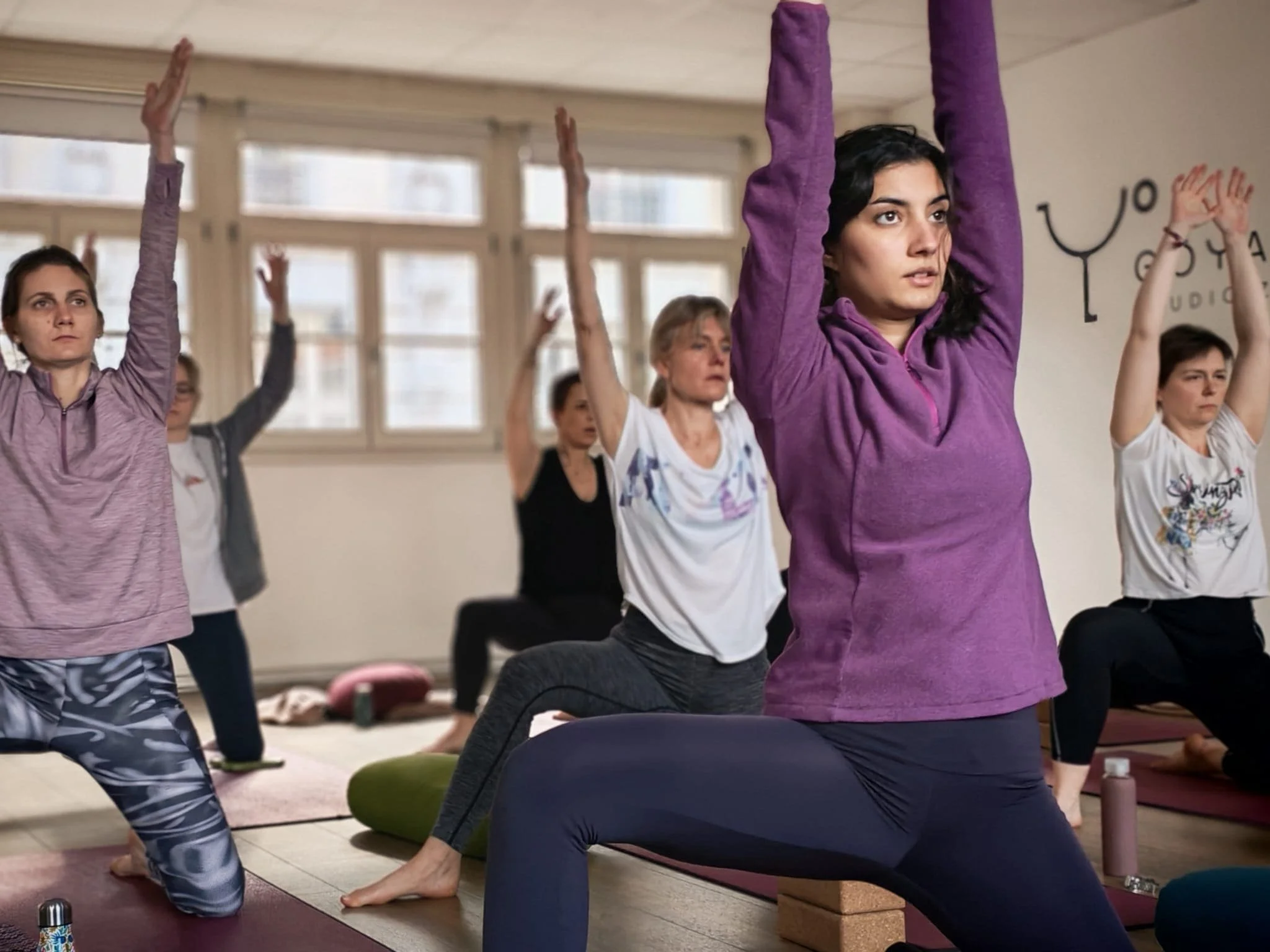 Groupe de personnes faisant du yoga en intérieur, en position de guerrier, avec des exercices de respiration, sur des tapis de yoga, dans une ambiance calme.